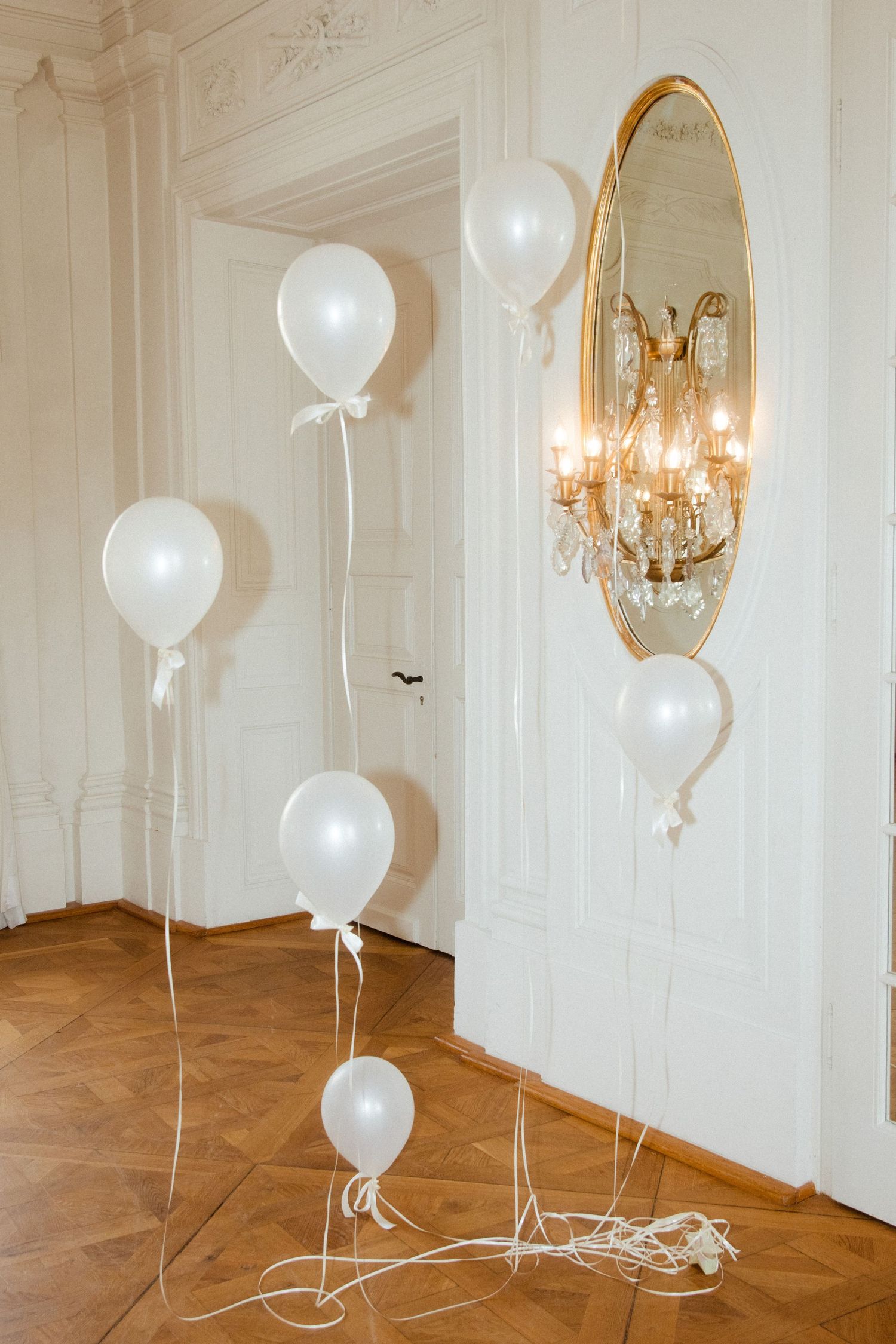 White helium balloons decorating a grand room with herringbone flooring and gilded mirrors.