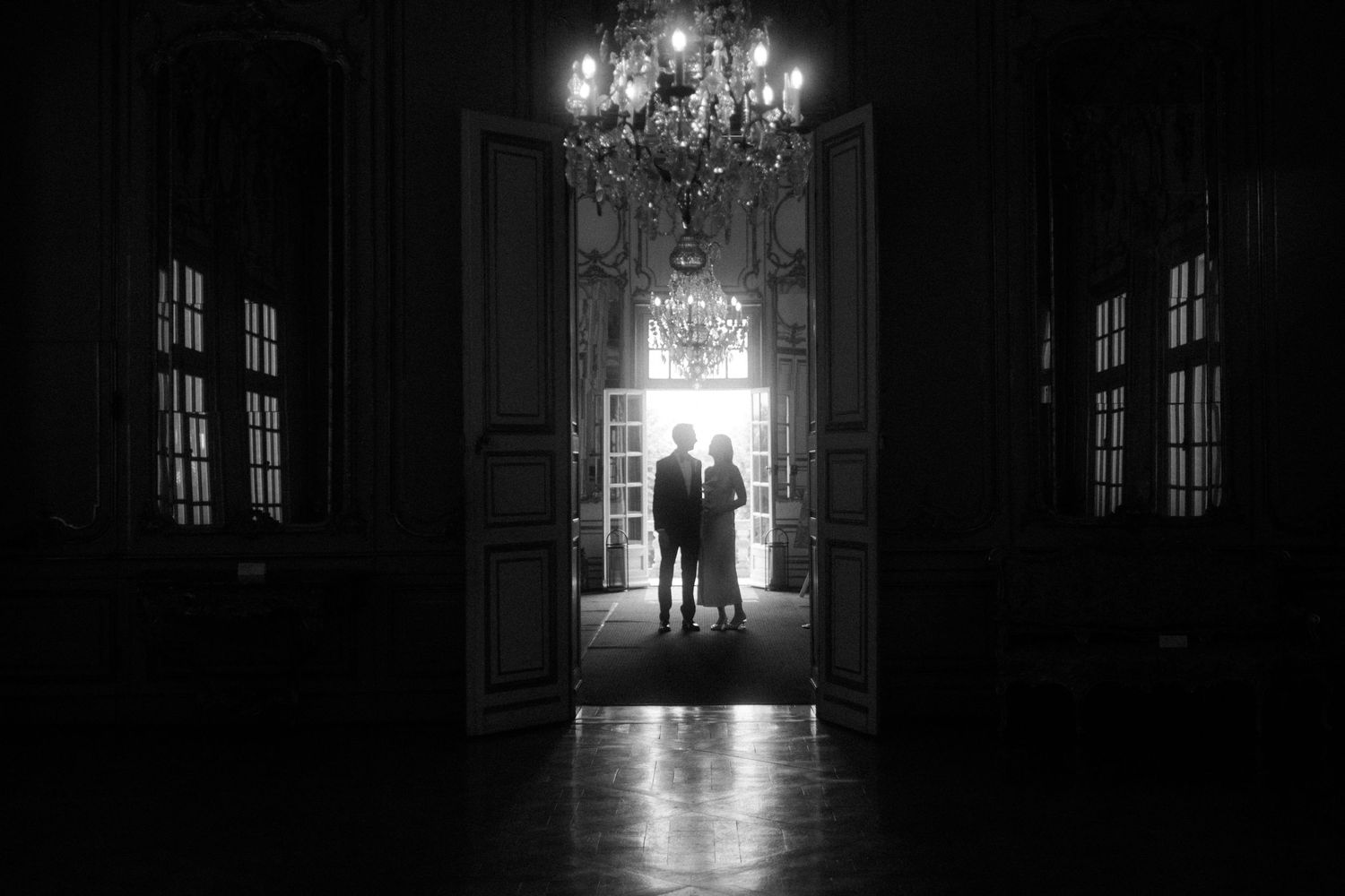 Silhouetted figures stand in a doorway of an elegant room with chandelier.