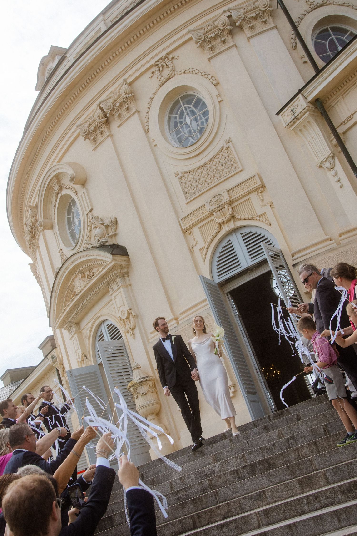 Wedding guests throw confetti as the couple exits a grand historic building with ornate architecture.