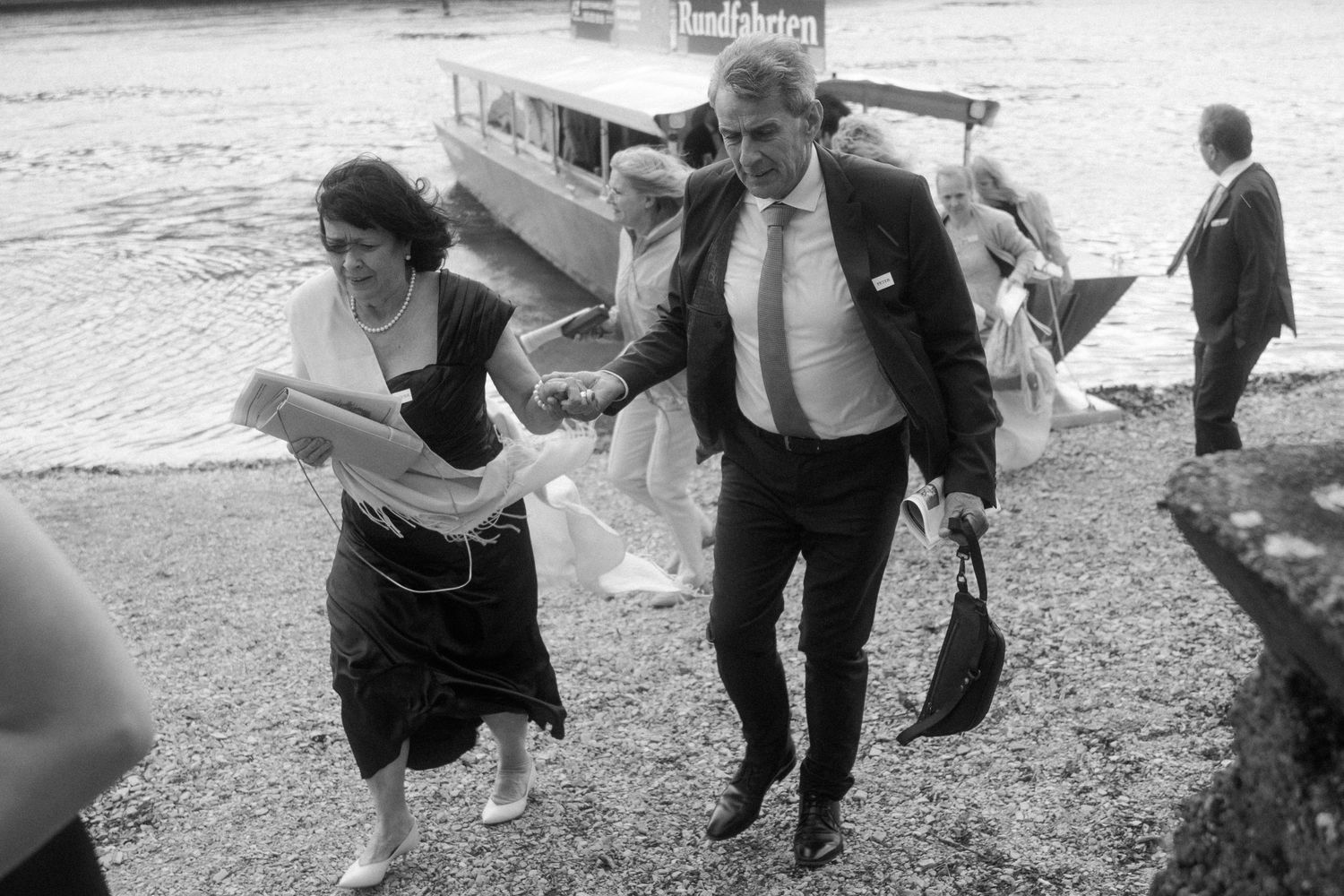 Black and white photo of a man and woman hurrying along a riverside path near a boat, dressed in formal attire.