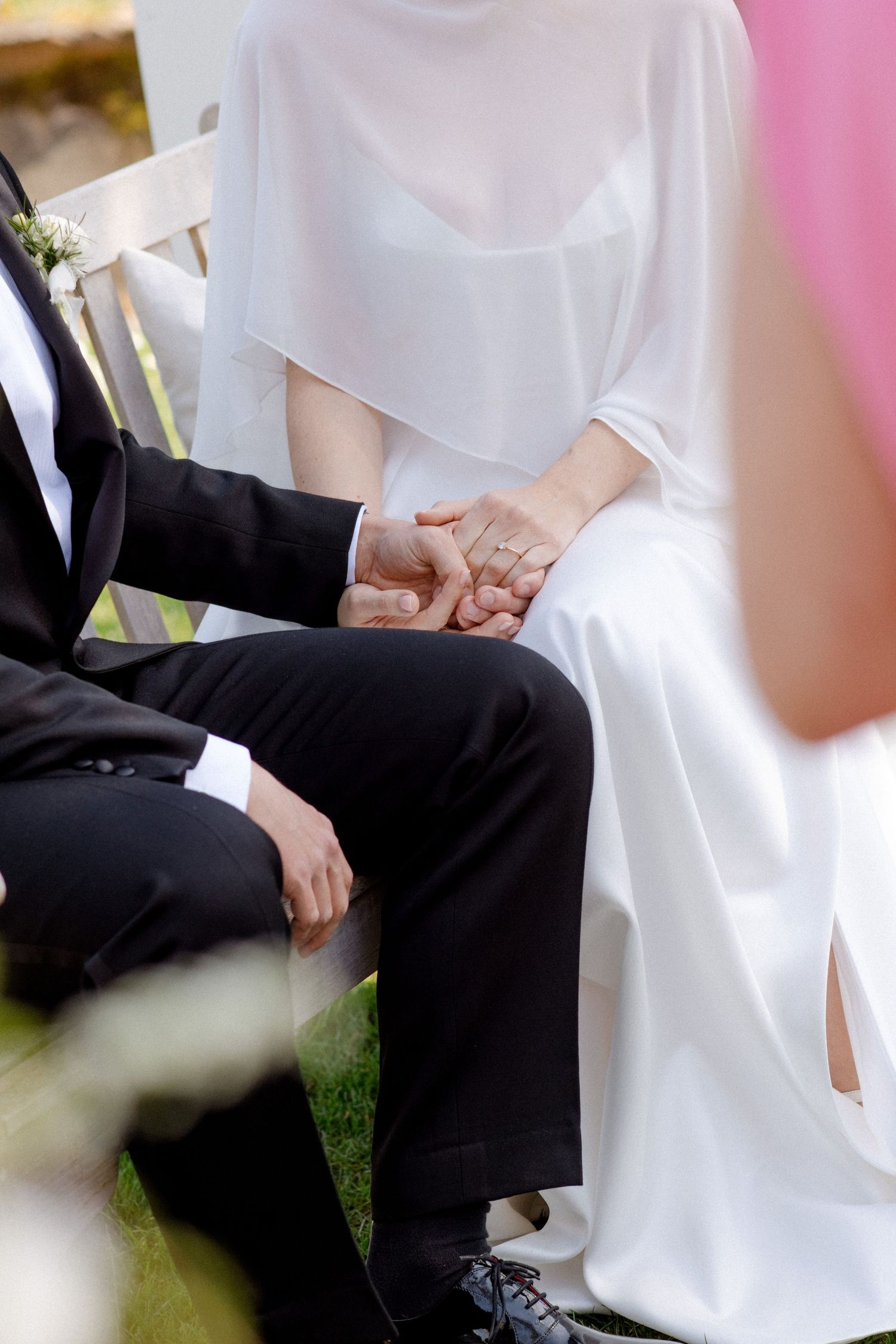 Bride in white dress and groom in black suit hold hands tenderly during outdoor wedding ceremony.