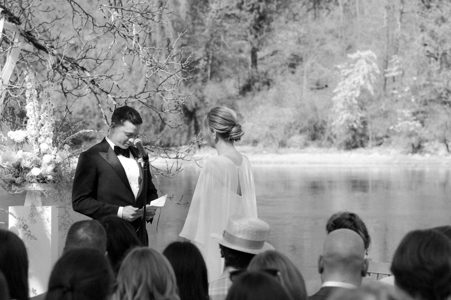 Black and white photo of a groom reading vows to his bride outdoors by a lake during a wedding ceremony.