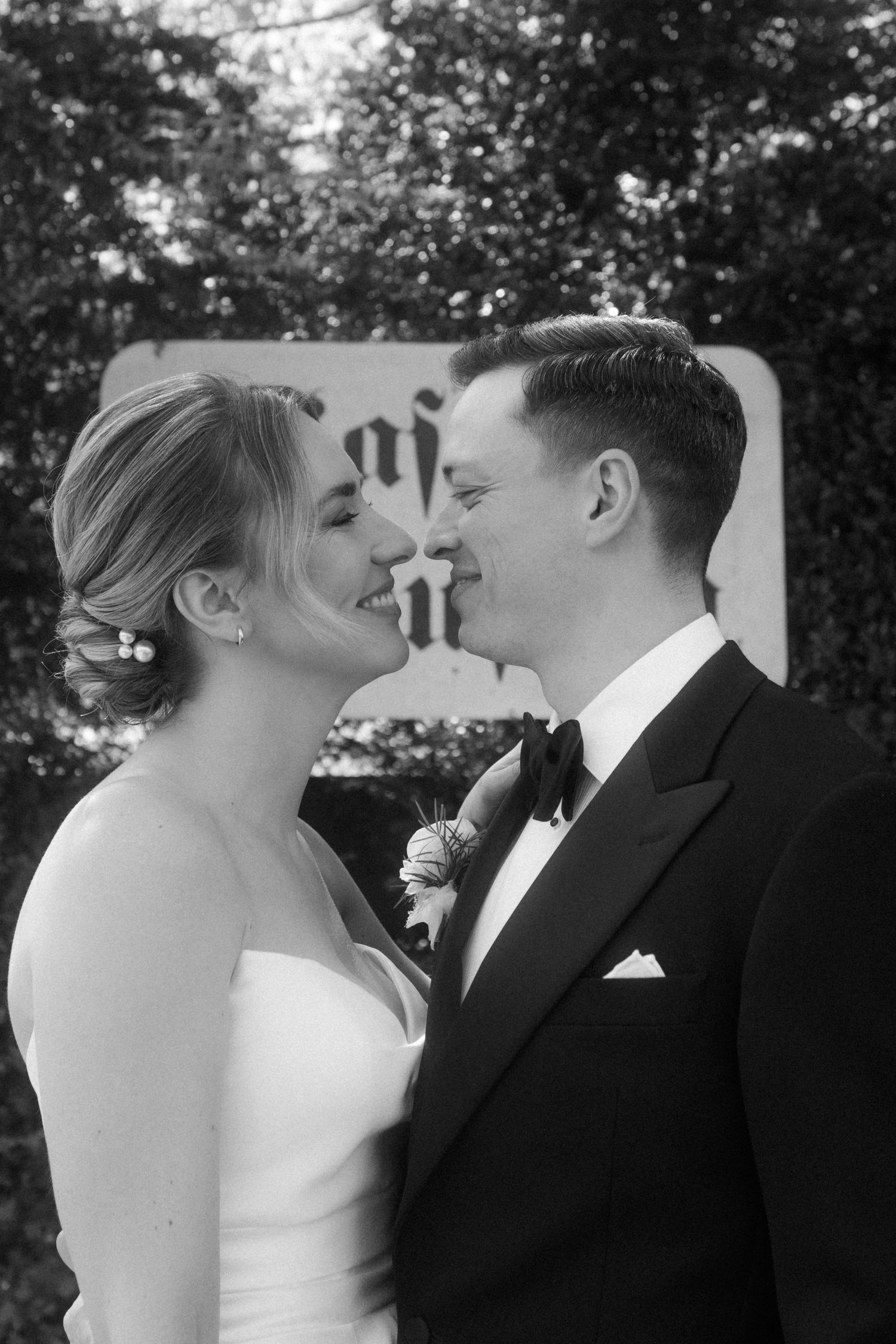 Black and white photo of a smiling bride and groom in formal wedding attire sharing a romantic moment outdoors.