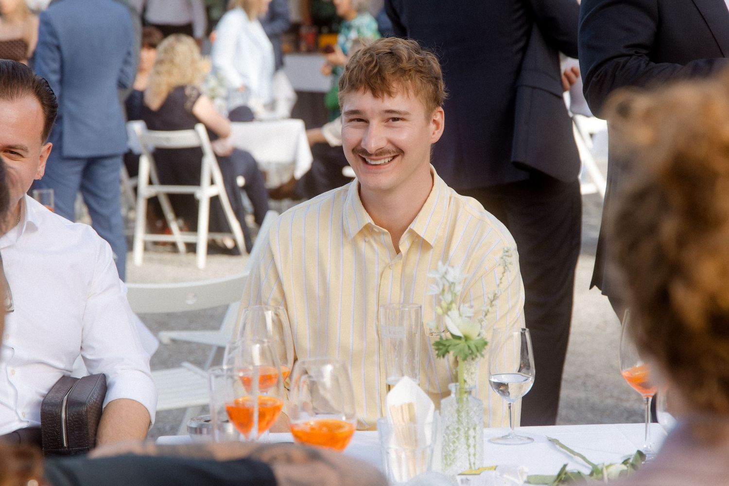 Young man with curly hair smiling at outdoor wedding reception dinner table.