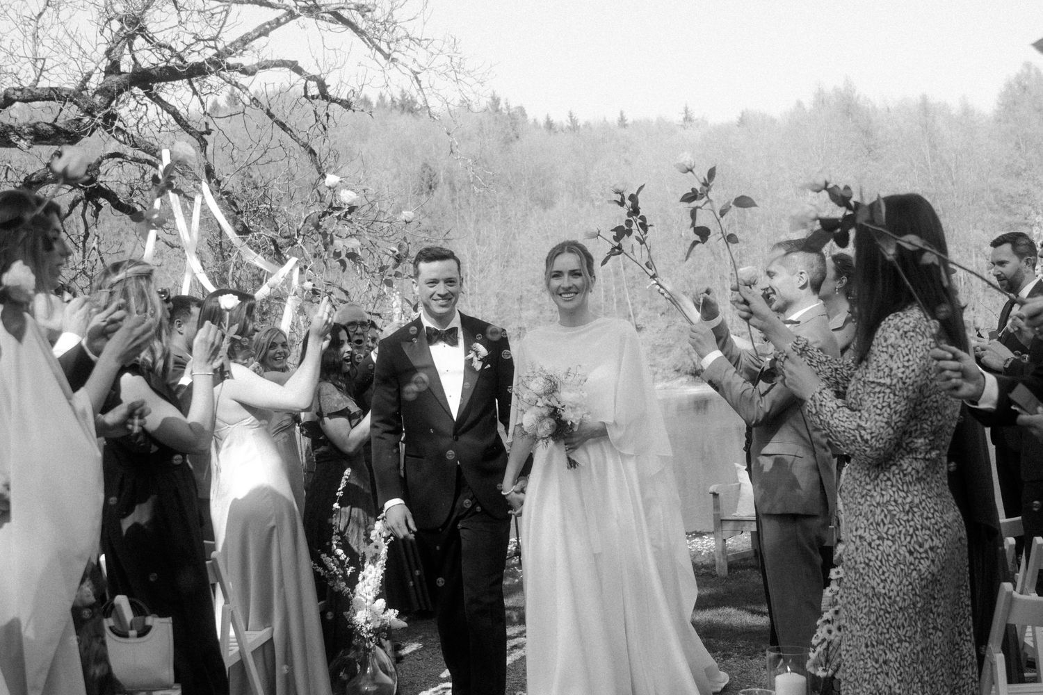 Black and white wedding photo of bride and groom walking through cheering guests outdoors.