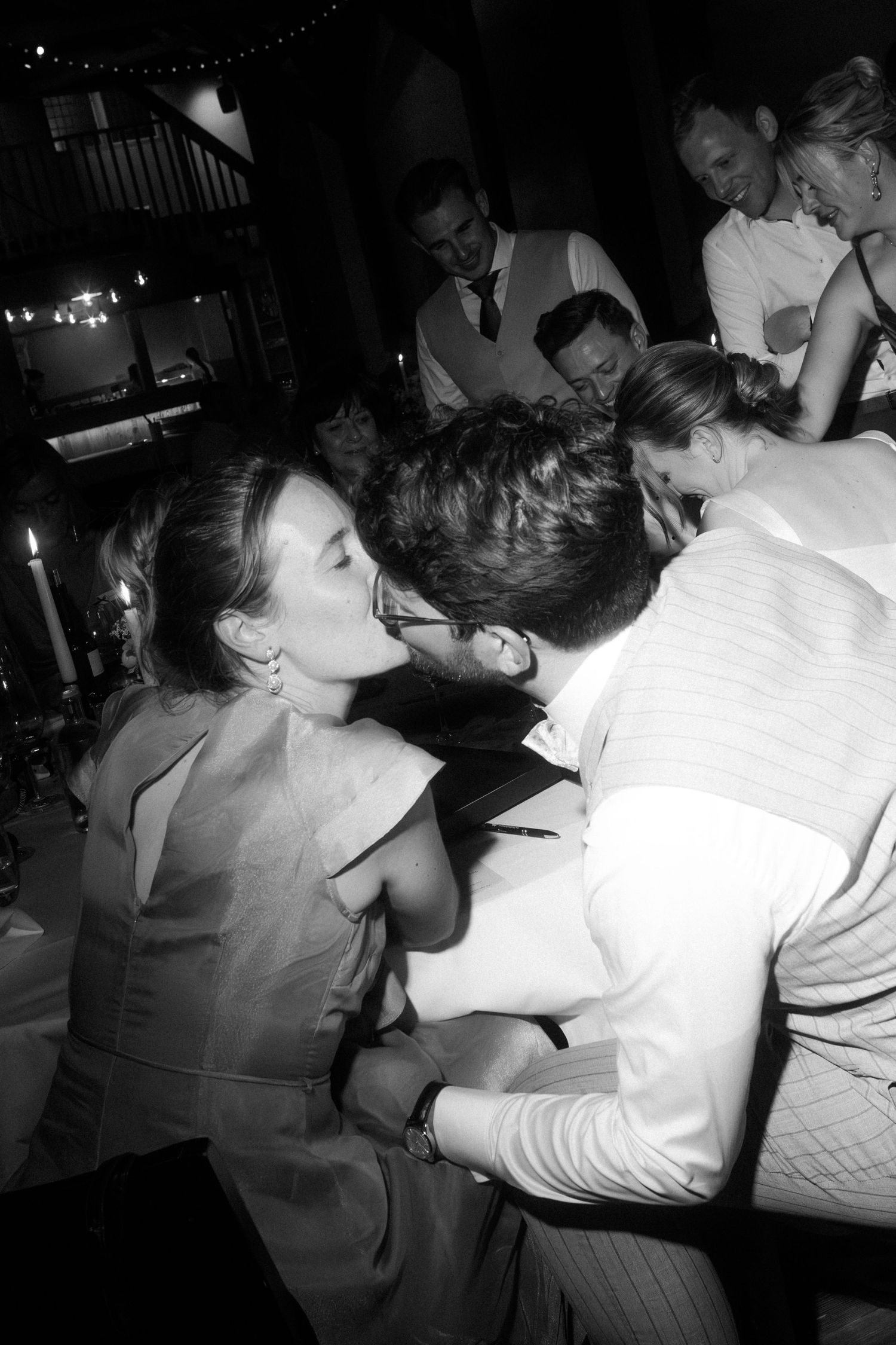 Black and white photo of couple sharing a kiss at a candlelit wedding reception surrounded by guests.