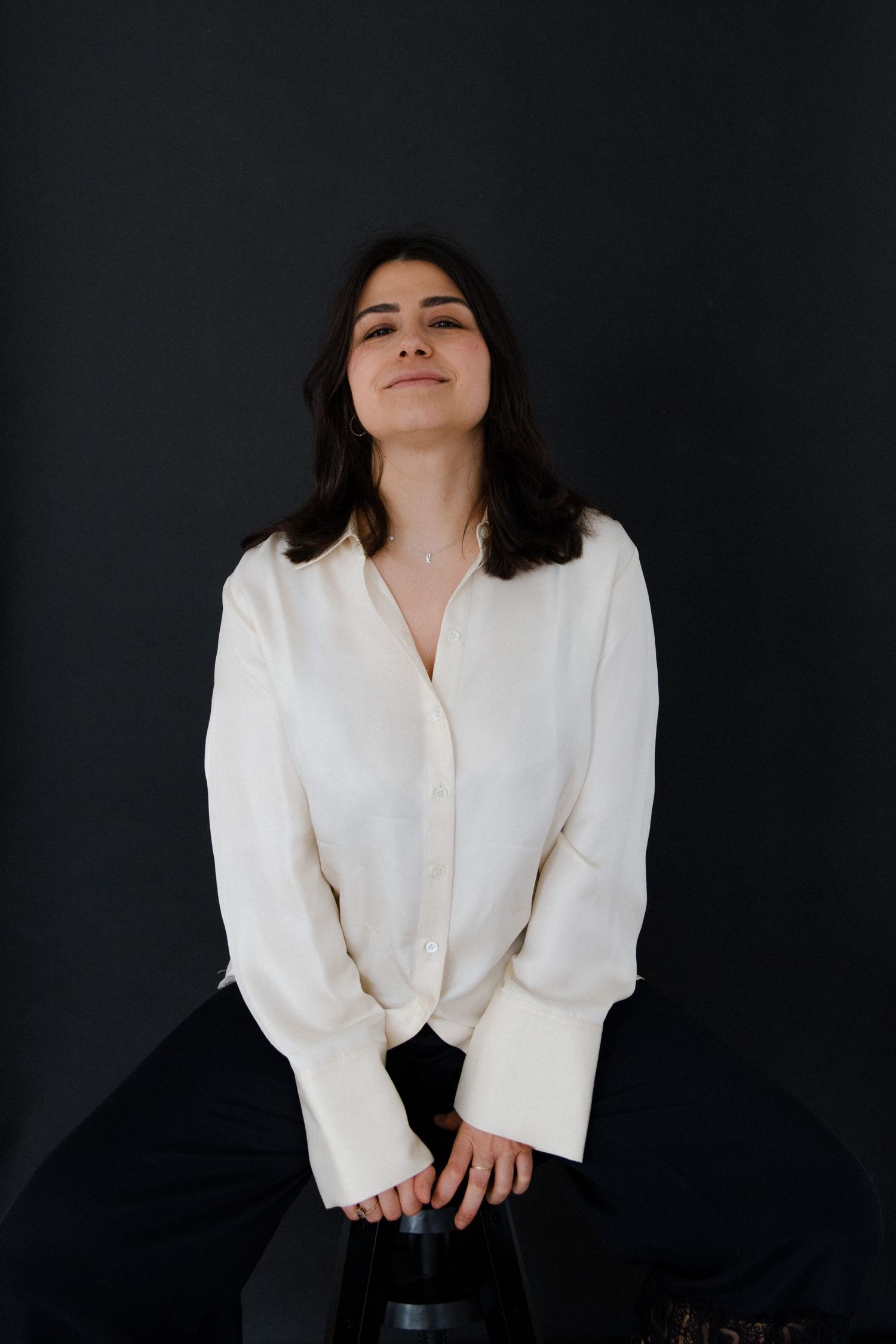 Young woman in white linen blouse seated against dark background, smiling softly with eyes slightly closed.