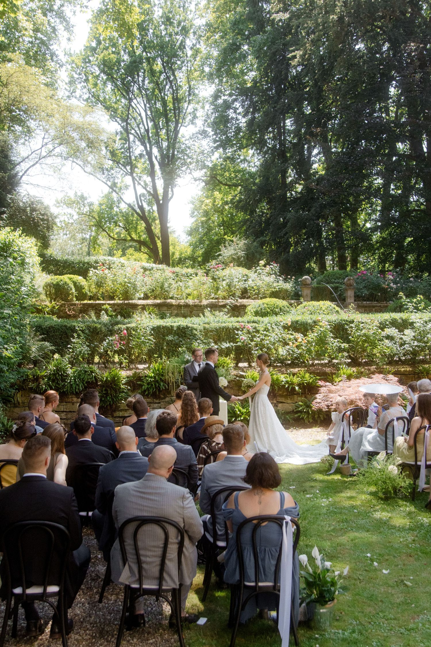 Outdoor garden wedding ceremony with guests seated on chairs facing the bride and groom beneath lush green trees.