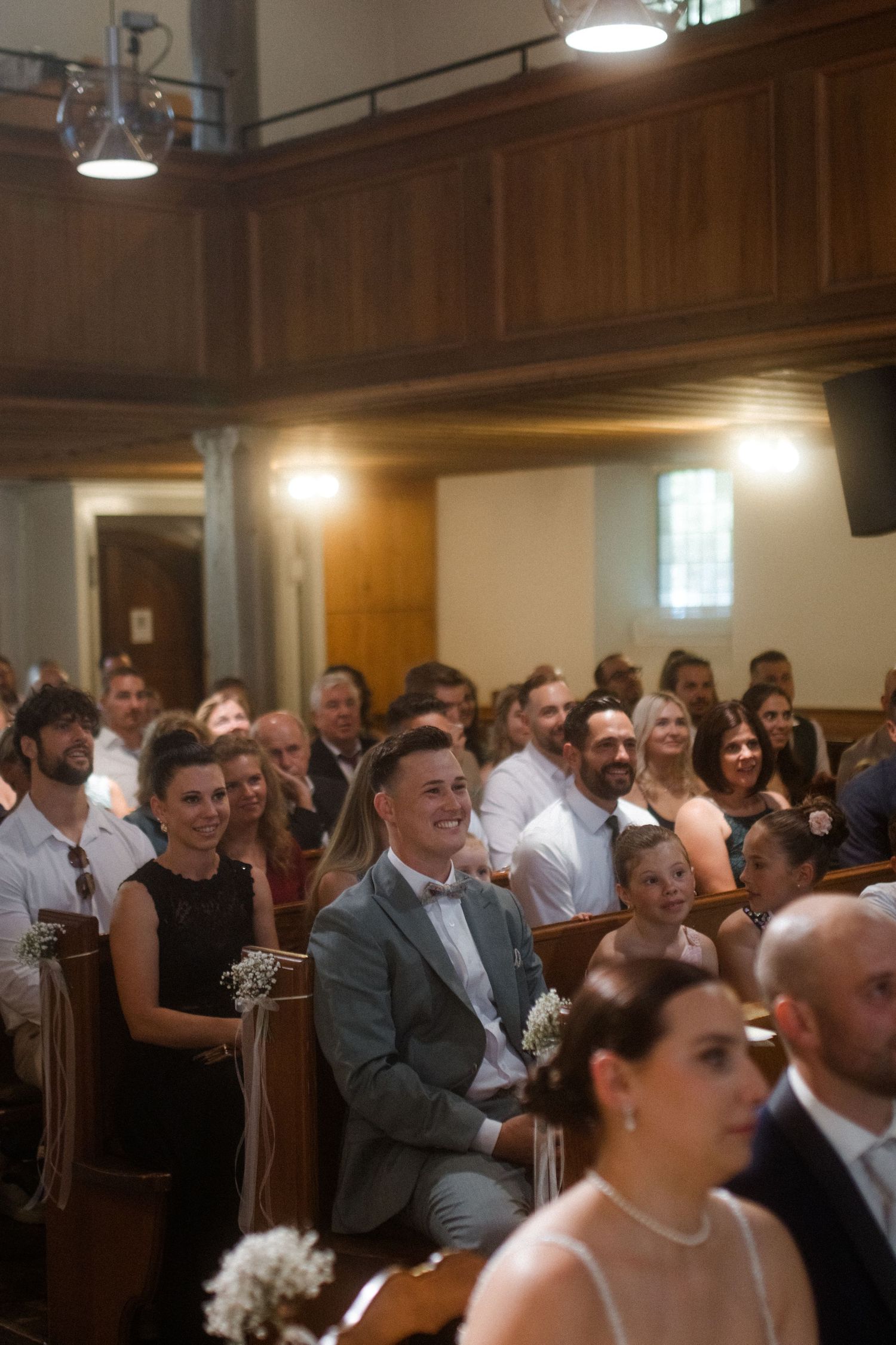 Wedding guests seated in wooden church pews with warm lighting and wood paneled walls.