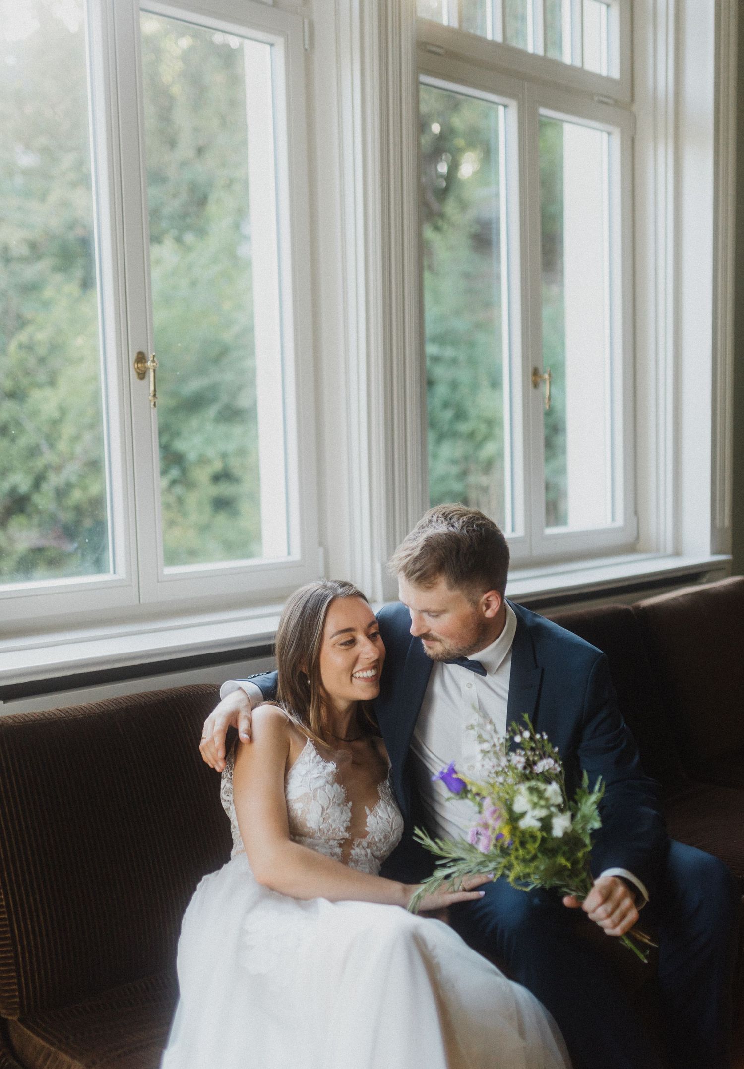 Elegant wedding couple sitting by sunlit windows in intimate moment.