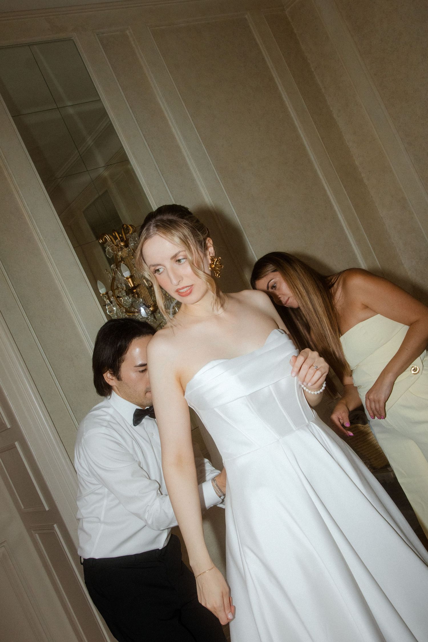 People helping with a white wedding dress fitting in an elegant room with chandelier.
