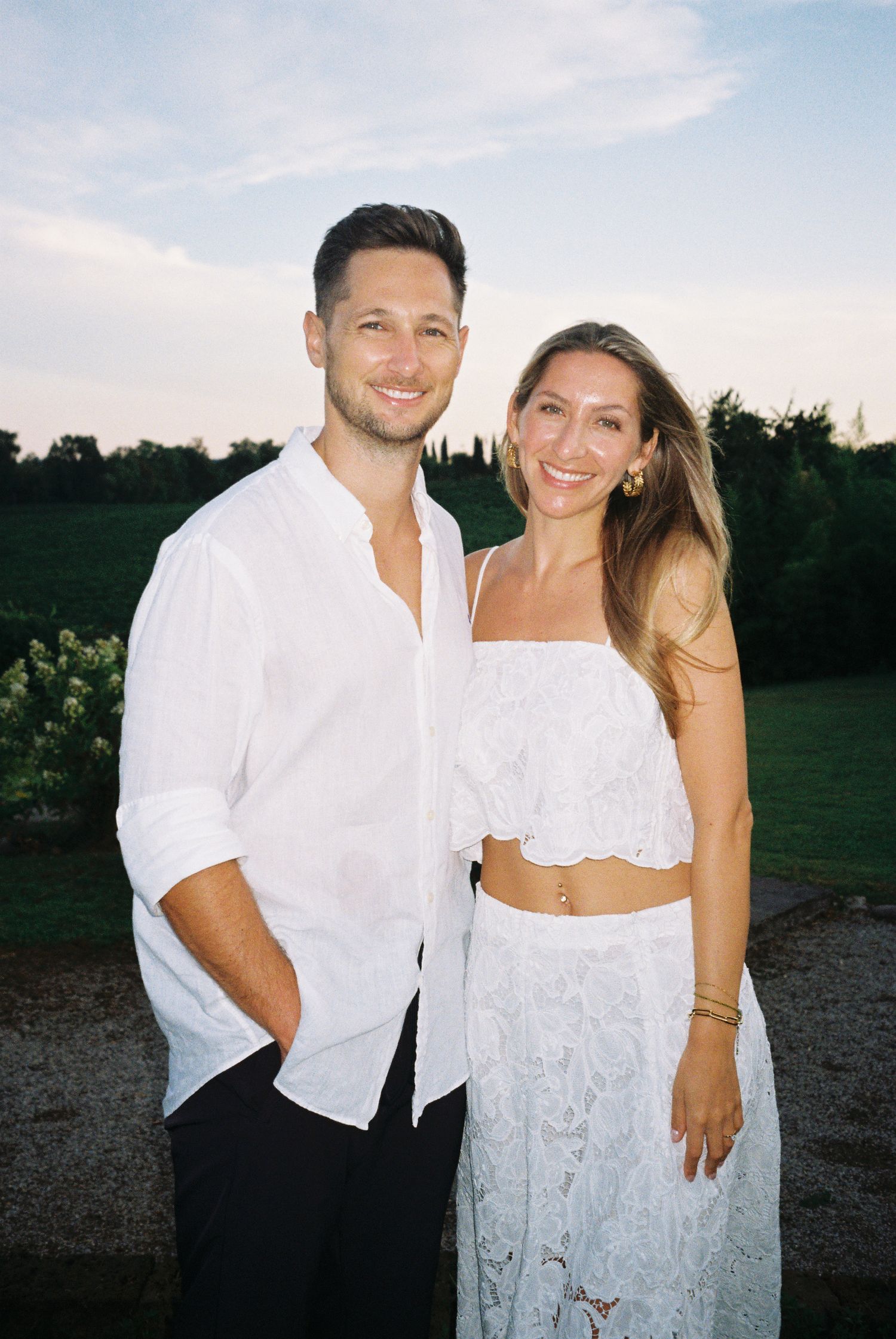 Couple in matching white outfits posing together at an outdoor summer event.