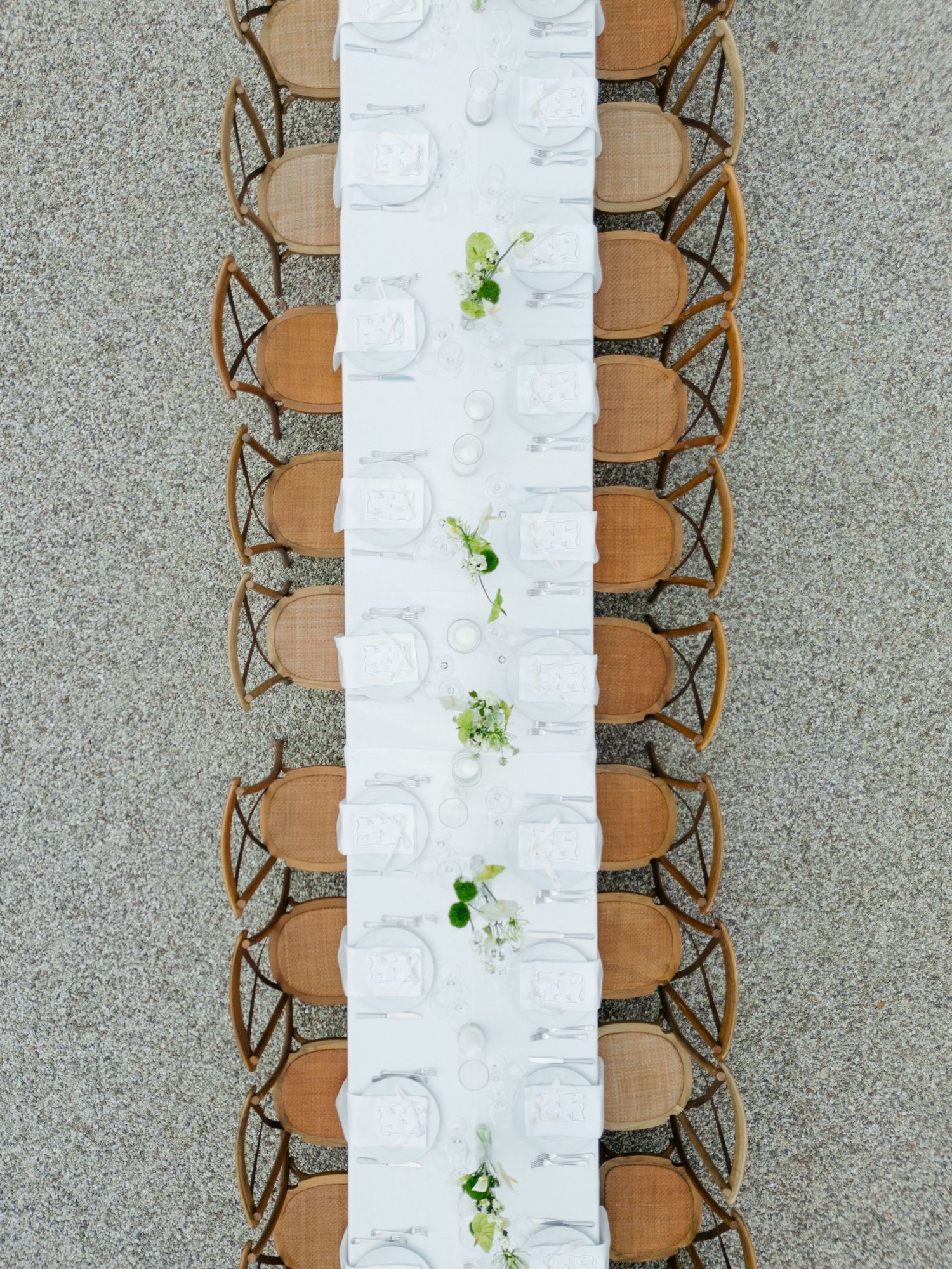 Overhead view of long dining table with white linens and cross-back chairs on stone patio.