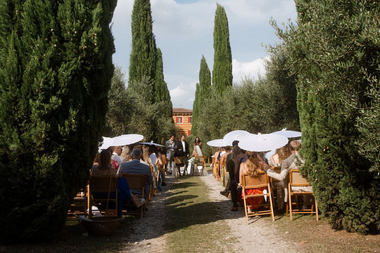 Outdoor wedding ceremony setup with cypress trees lining a path and guests seated under white parasols.
