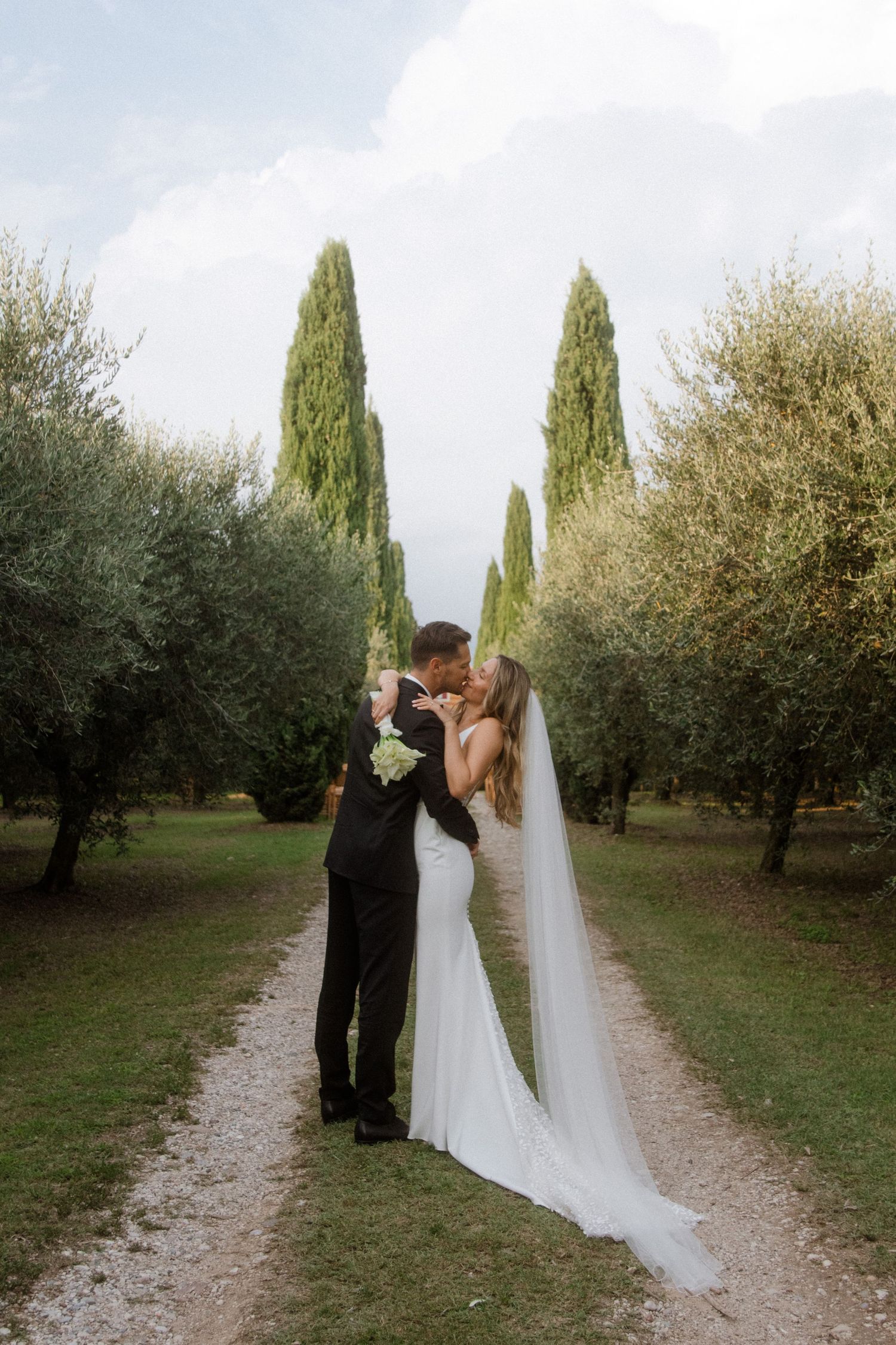 Bride and groom kissing on a tree-lined path in Tuscany, bride wearing a white gown with long veil.