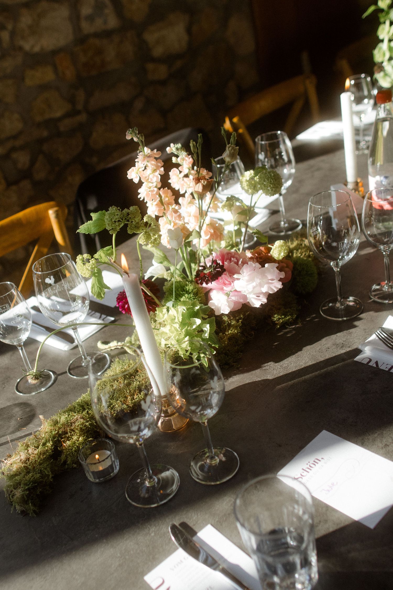 Elegant table setting with wine glasses, moss decor, and floral centerpieces against stone wall.