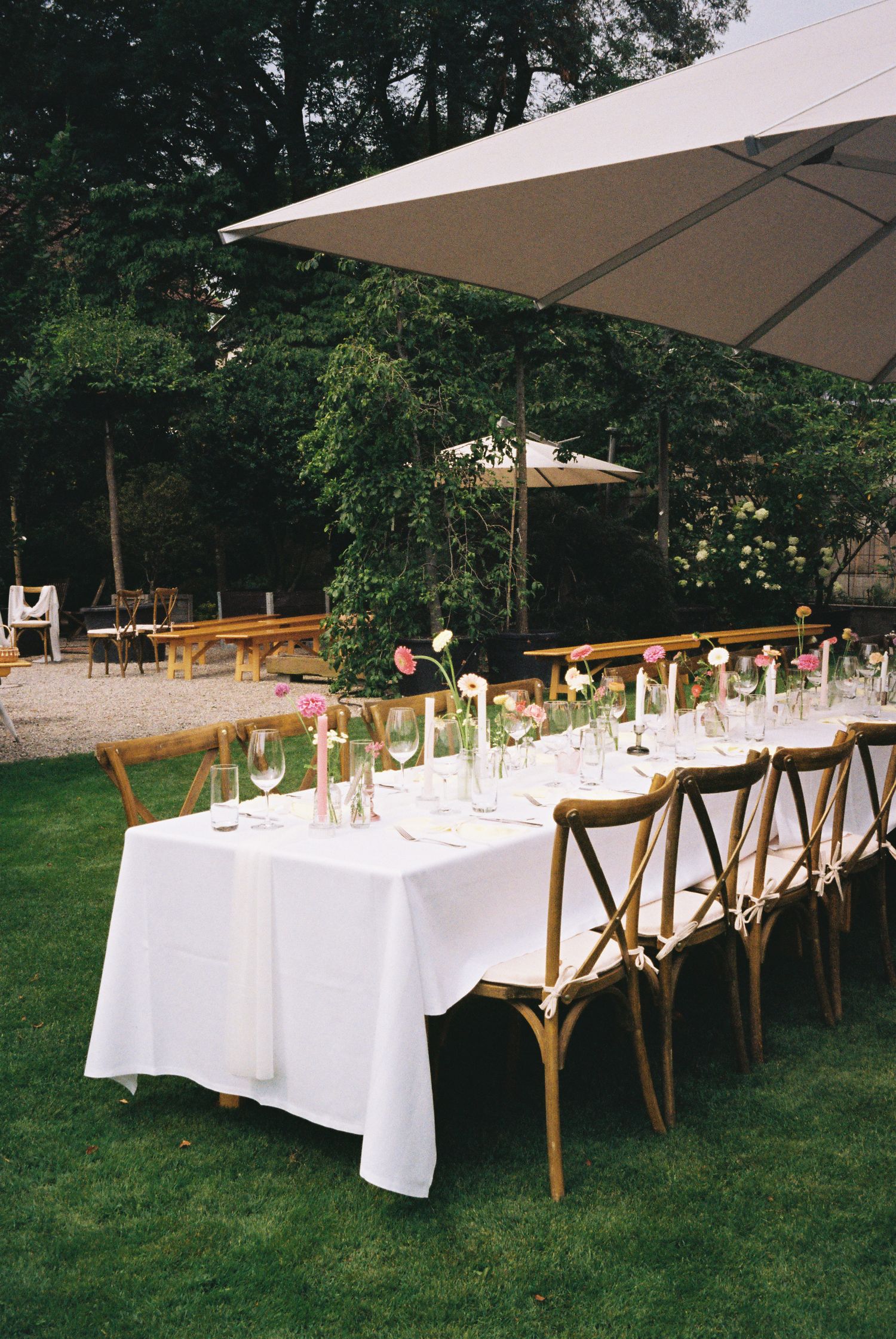 Long white dining tables with wooden cross-back chairs set up outdoors under beige umbrellas for an elegant garden party.