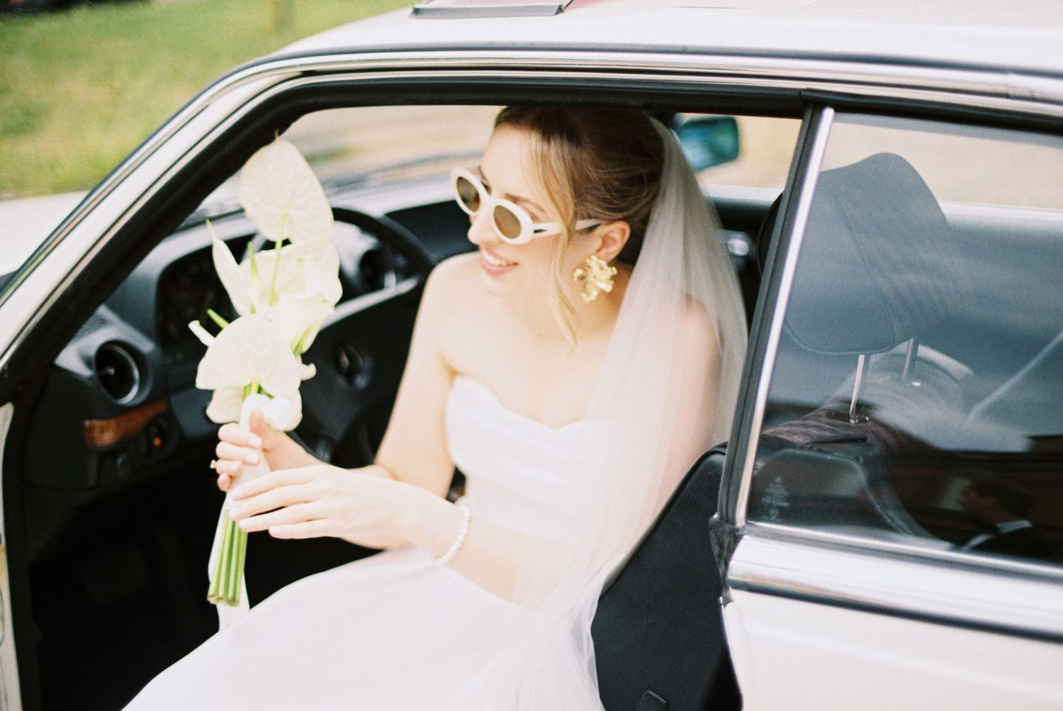 A bride in white dress and sunglasses holds a bouquet of white flowers while seated in a car.