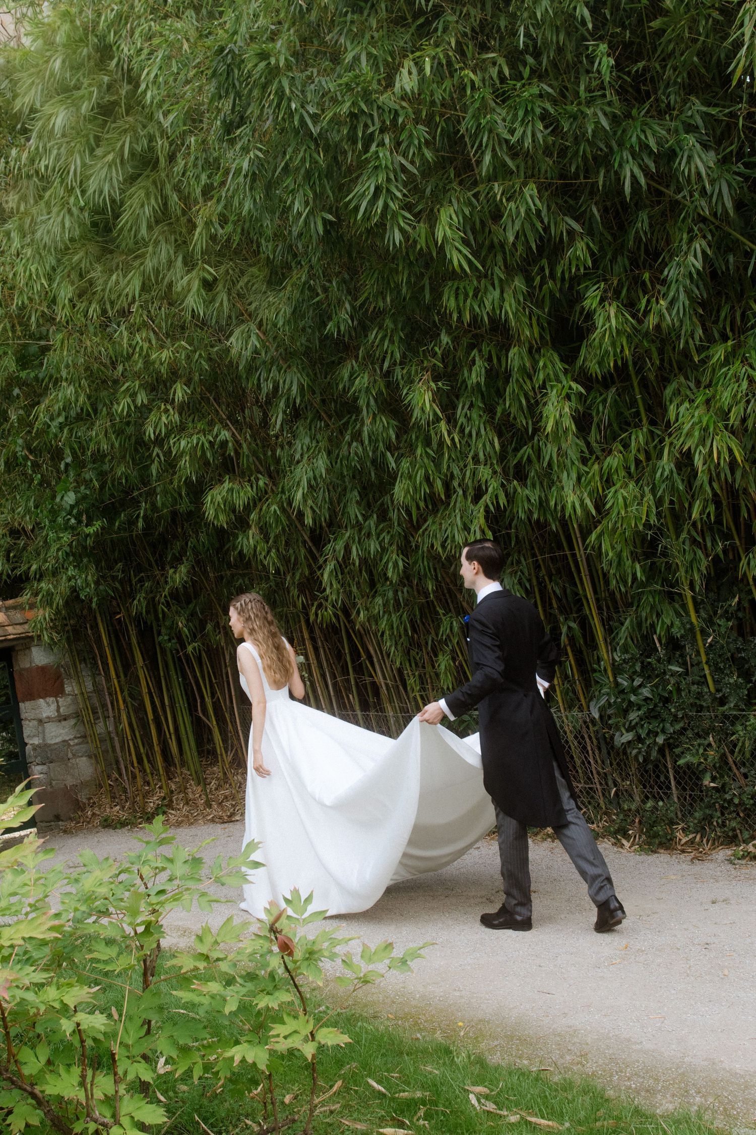 A couple in wedding attire walks along a garden path lined with bamboo plants.