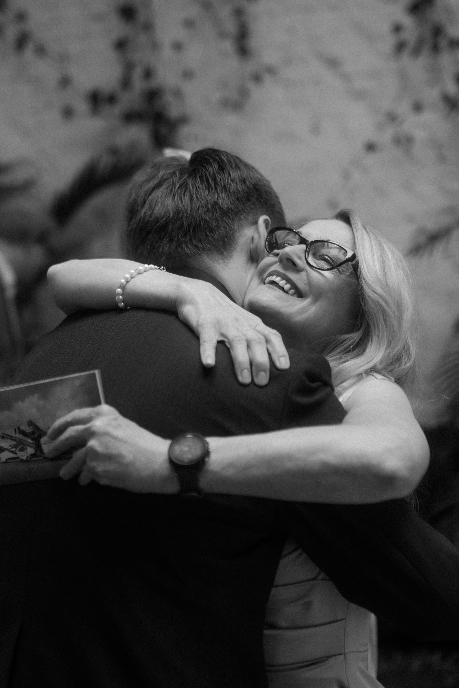 A black and white photo captures an intimate moment of laughter and hugging at a celebration.
