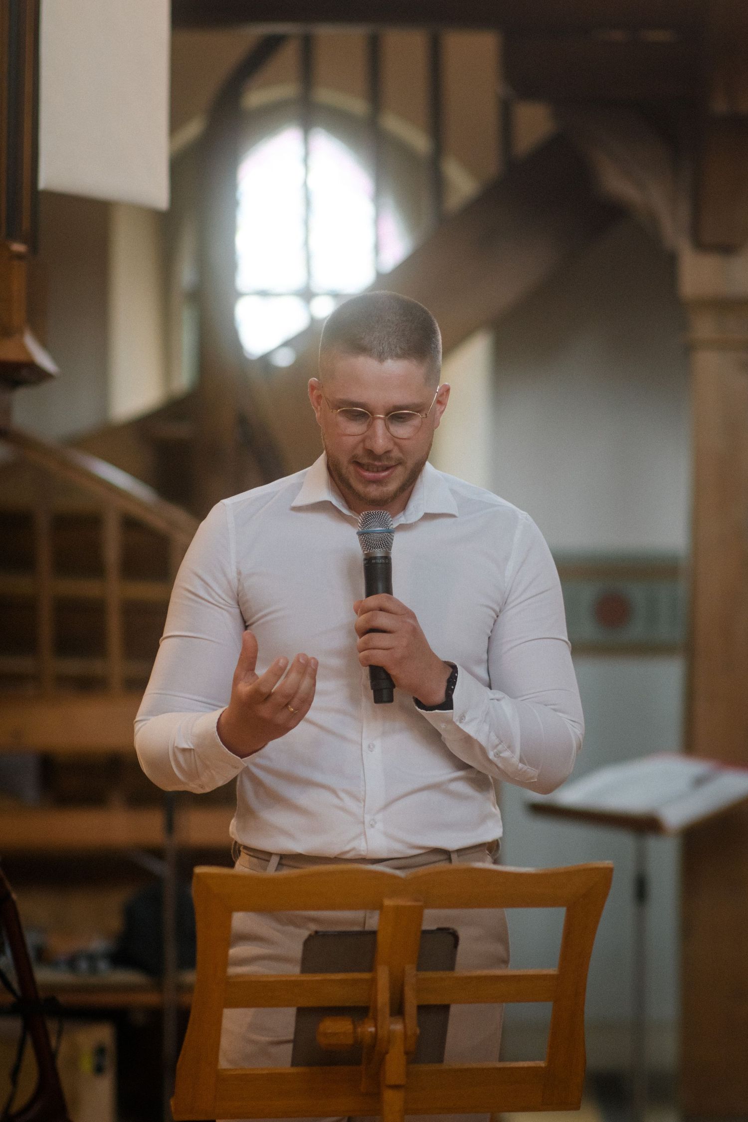 A speaker in a white dress shirt delivers a message while standing in a church setting with a wooden chair behind him.