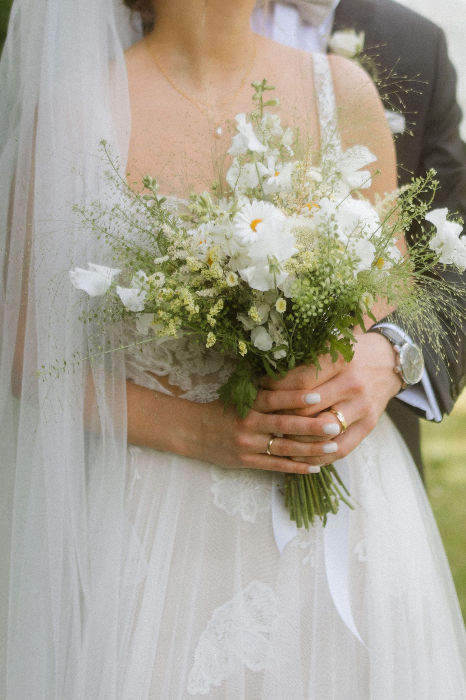 Close-up of a delicate white and green wedding bouquet held by bride in lace dress with flowing veil.