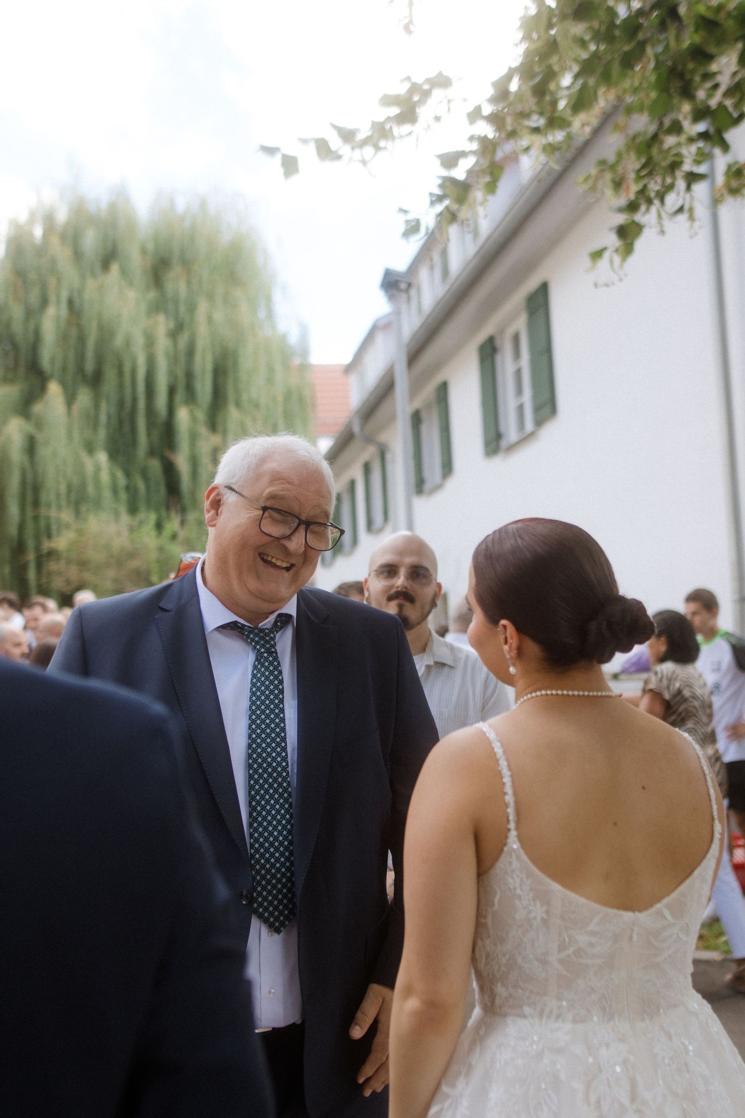 A bride in a white dress shares a joyful moment with an older gentleman wearing glasses and a blue suit at a wedding.