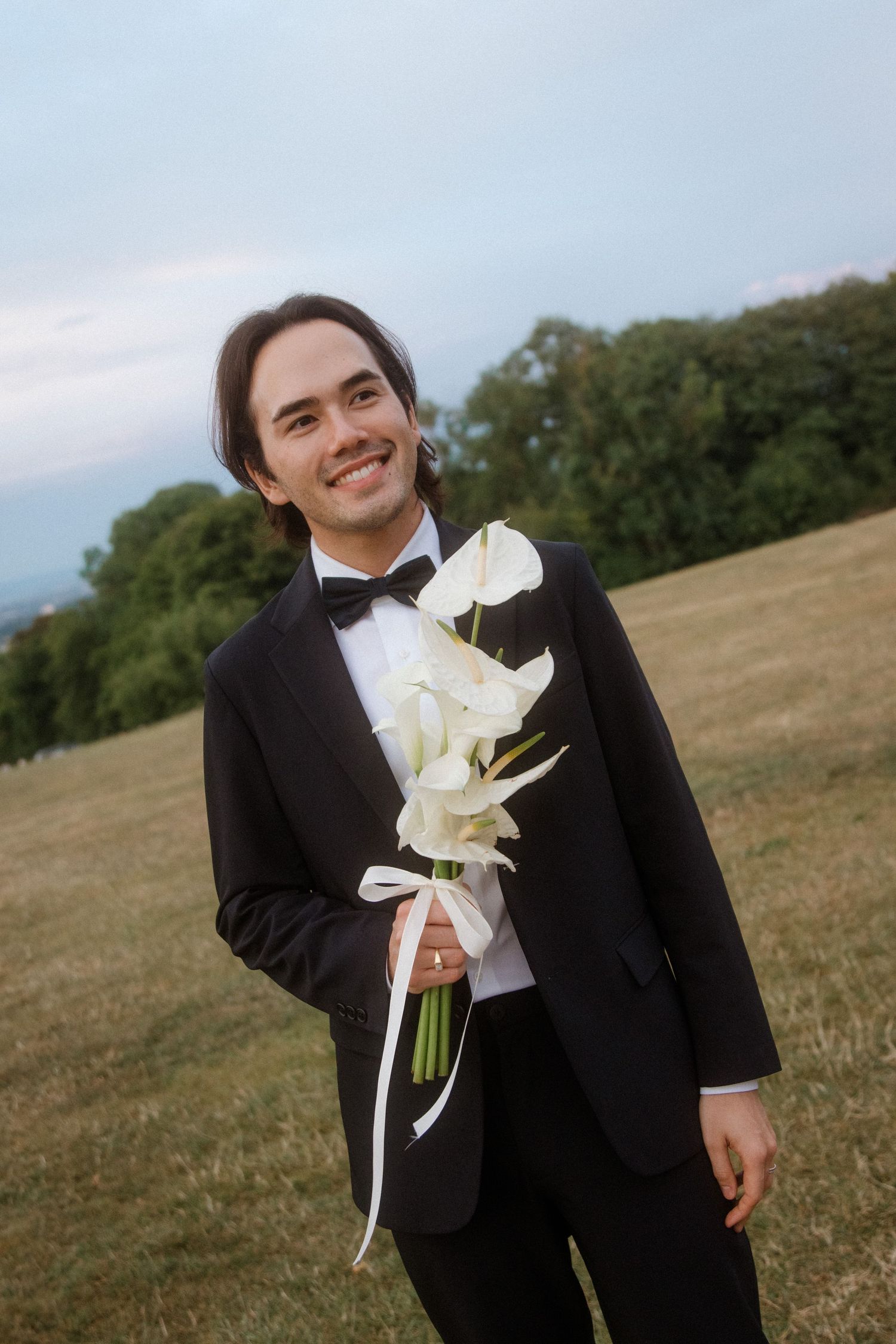 Person in black tuxedo holding white calla lily bouquet against grassy field.