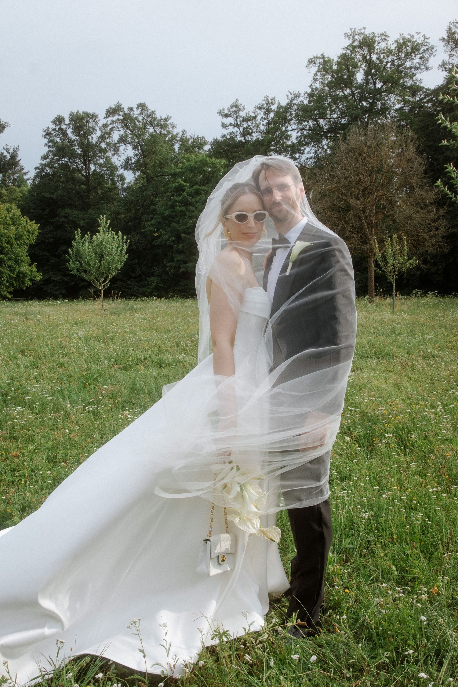 A romantic wedding couple poses in a meadow as the bride's veil flows dramatically in the wind.