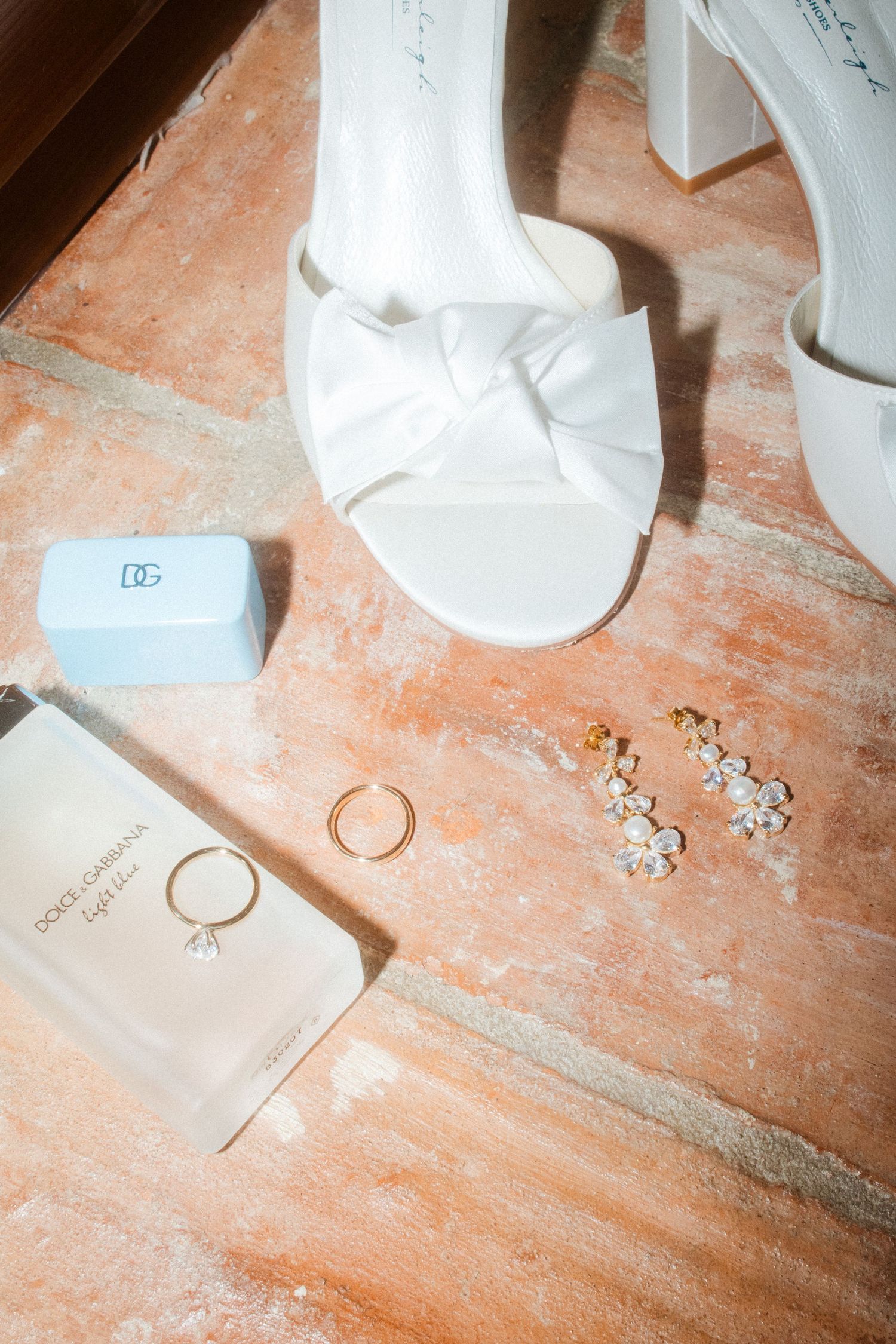 Wedding accessories including white bow shoes, rings, and earrings arranged on terra cotta flooring.