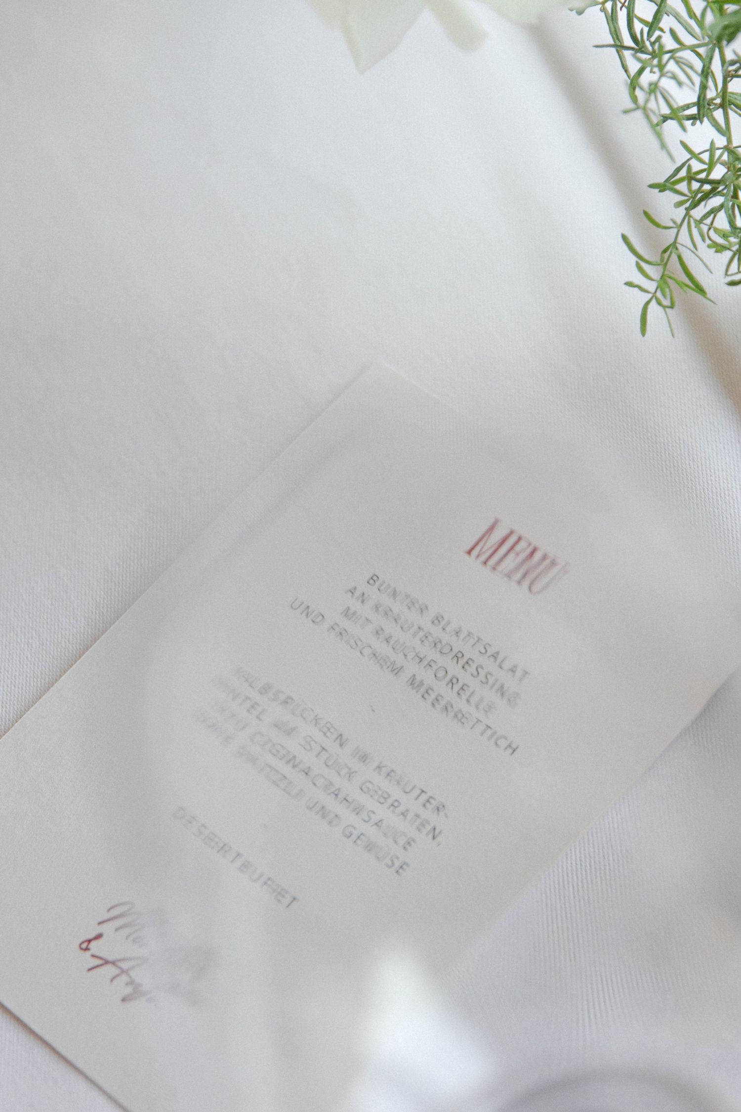 Elegant wedding menu card on white fabric with green herb sprig decoration.