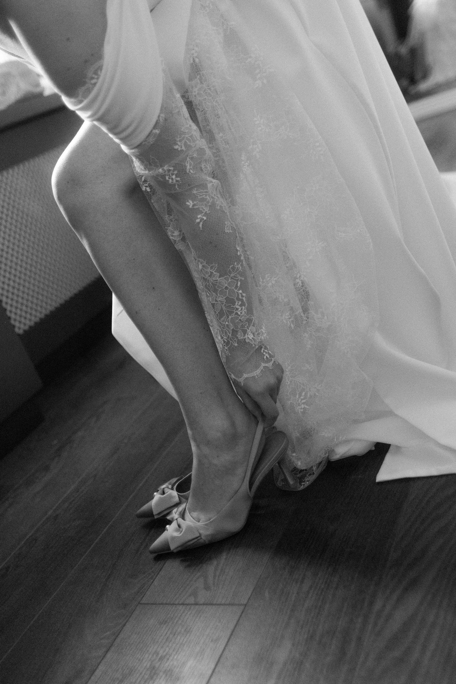 Bride in lace wedding dress putting on elegant heeled shoes with bow detail on hardwood floor, black and white photo.