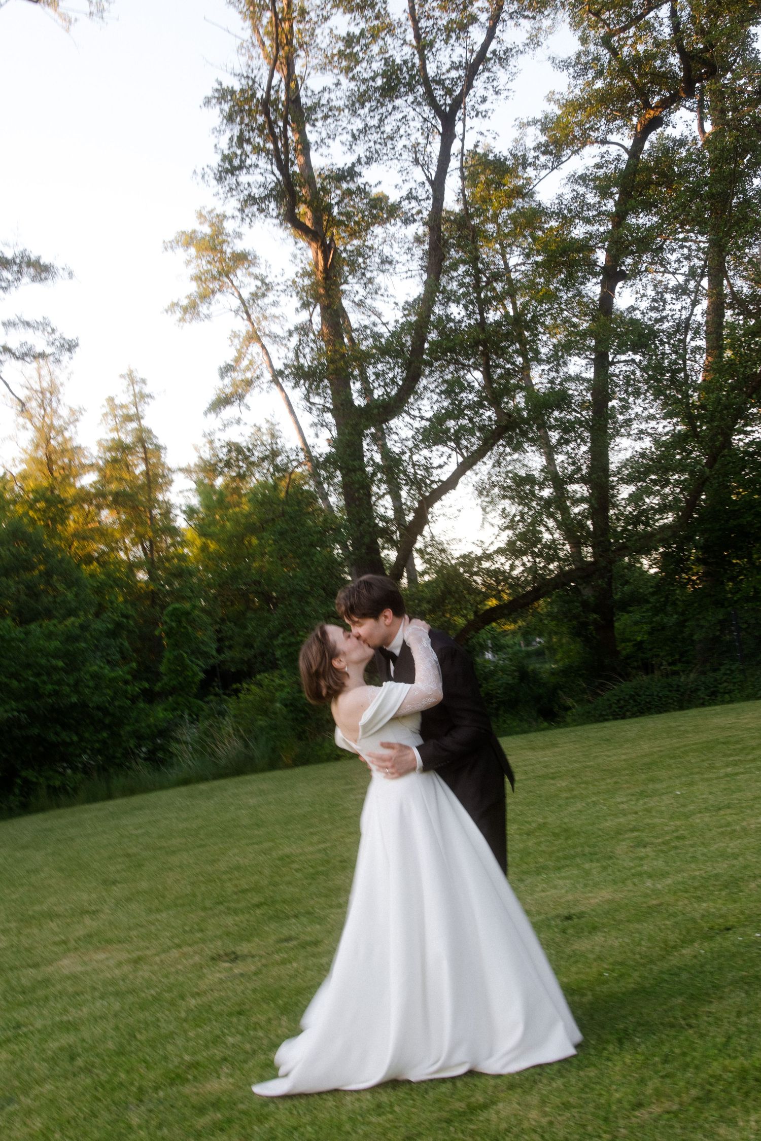 Bride and groom share a romantic kiss on a lush green lawn surrounded by trees at golden hour.