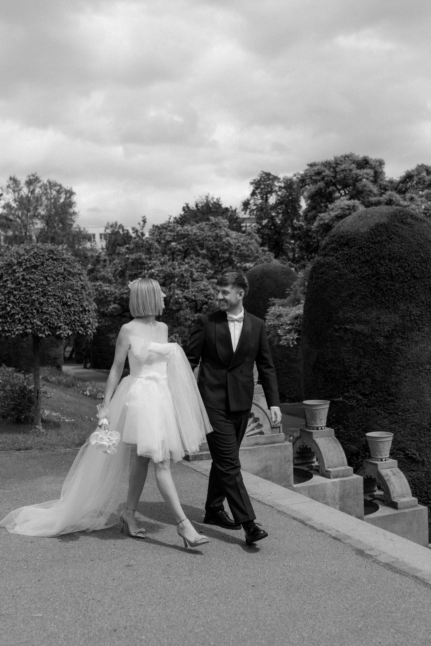 Black and white photo of a couple walking along a garden path in formal wedding attire.