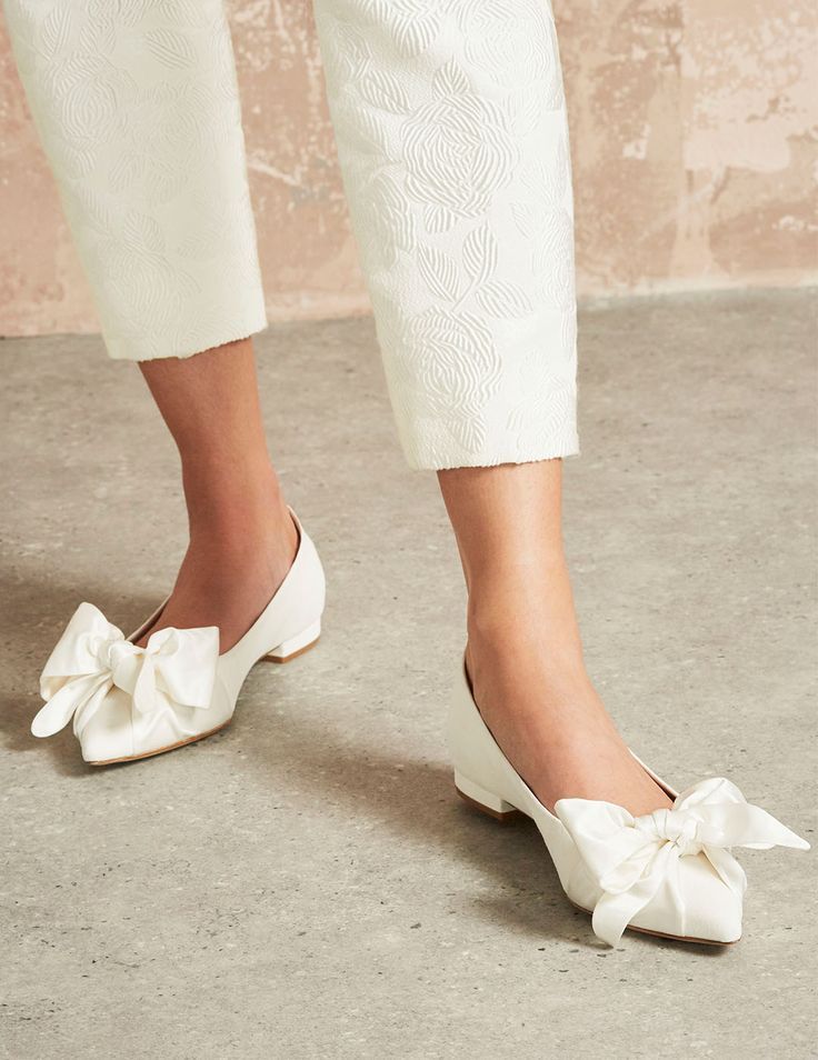 White bow-adorned flat shoes paired with cropped white pants on concrete floor.