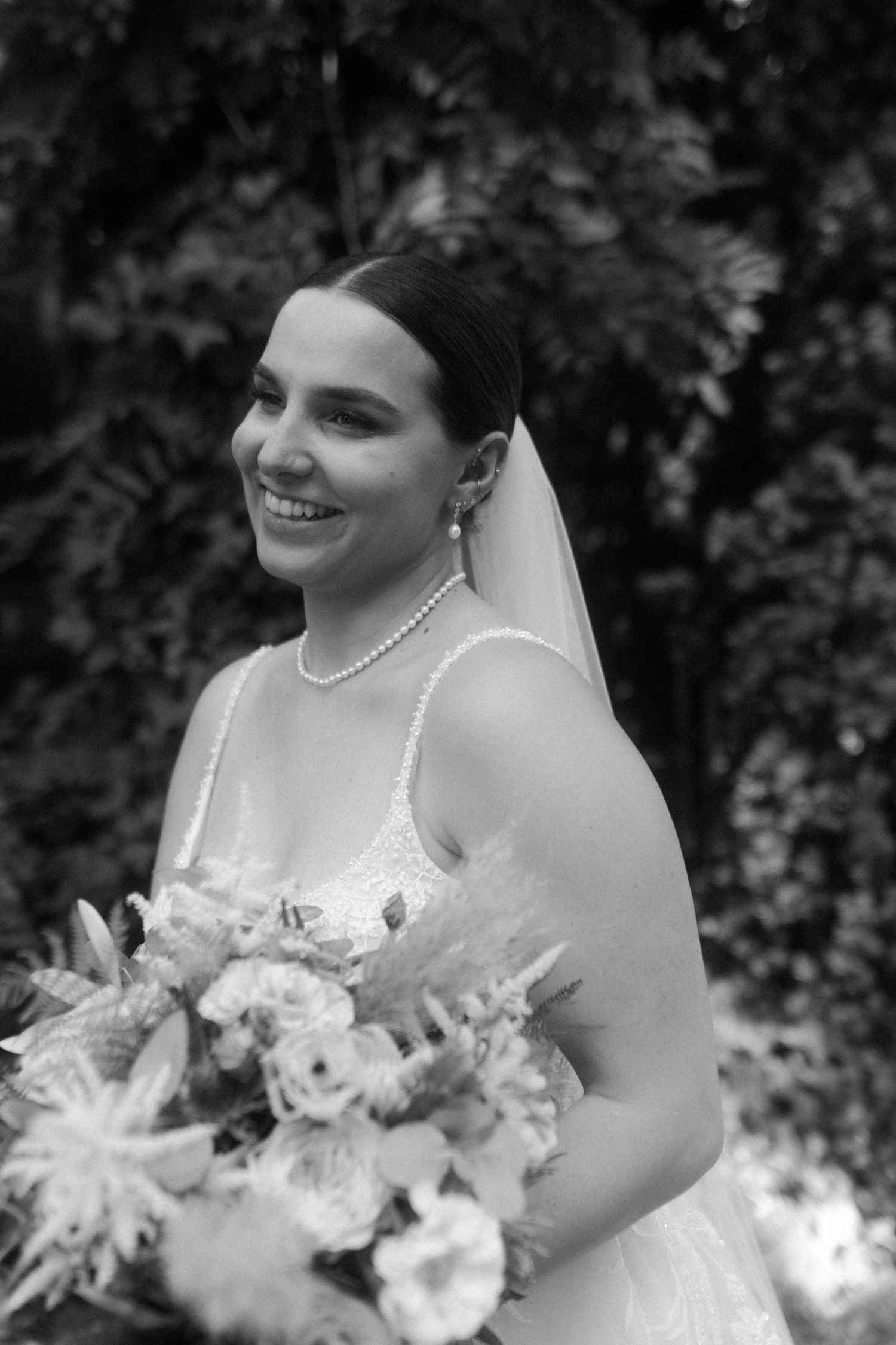 Black and white portrait of a smiling bride holding a lush bouquet with delicate lace dress details.