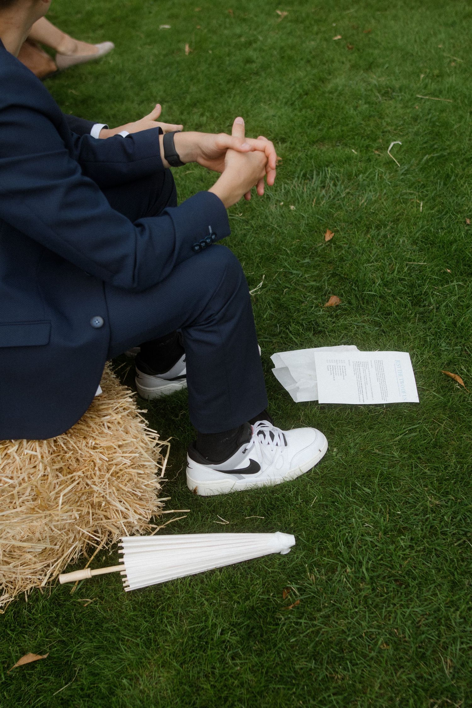 Person in navy suit sits on hay bale holding white parasol at outdoor event.