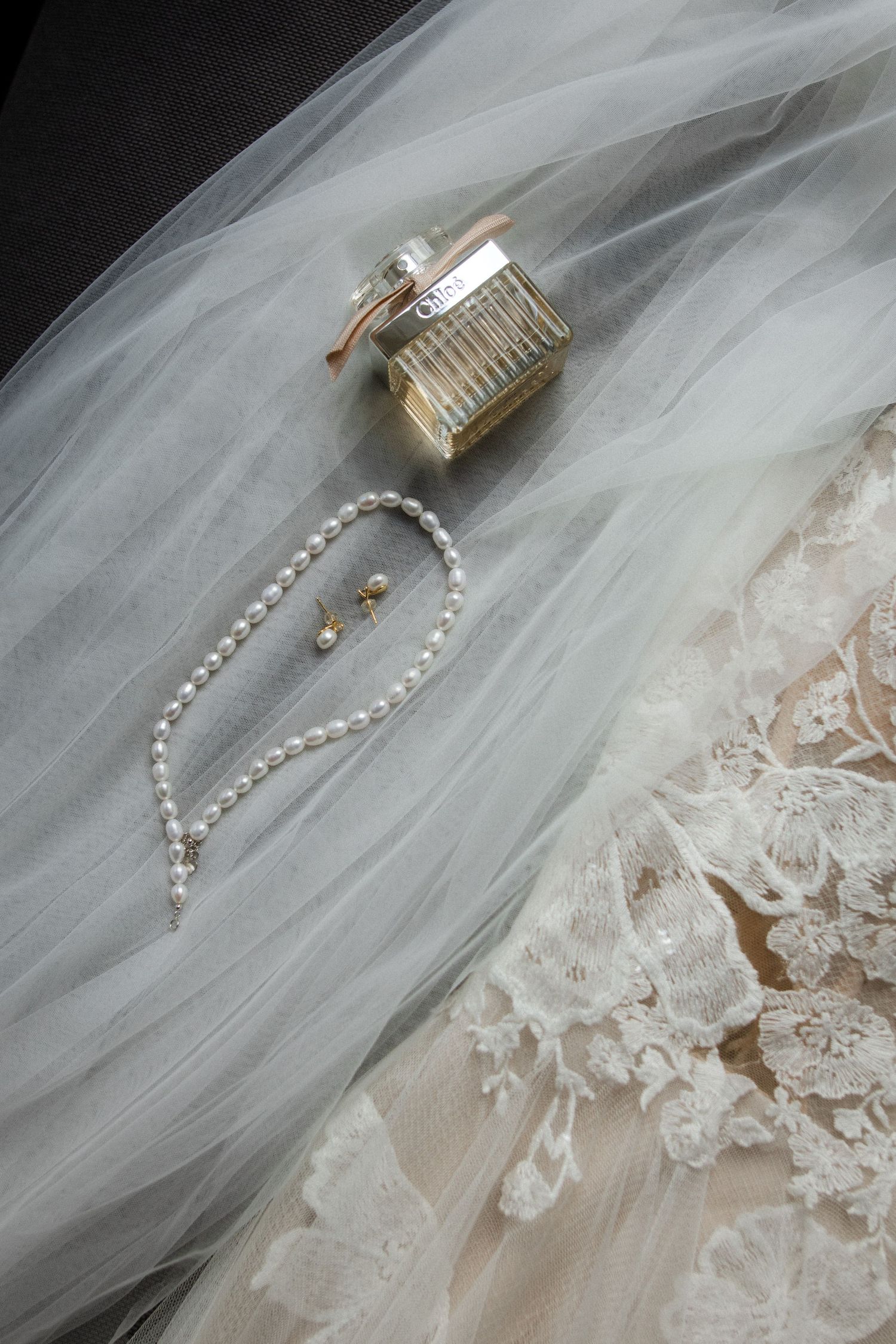 Close-up of bridal accessories including perfume bottle, pearl necklace and lace detail on tulle fabric.