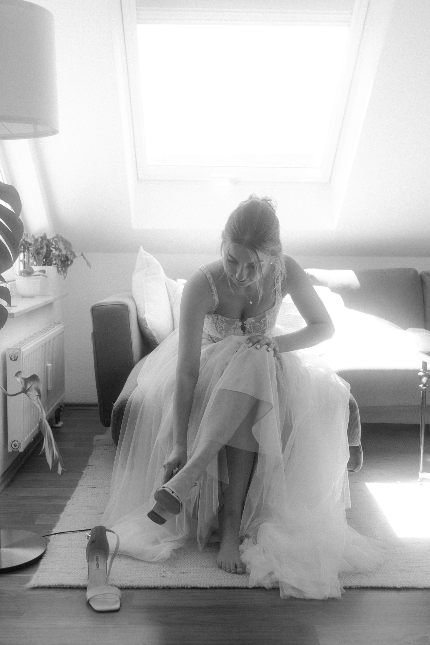 A bride in a flowing white gown sits in sunlight while putting on her wedding shoes.