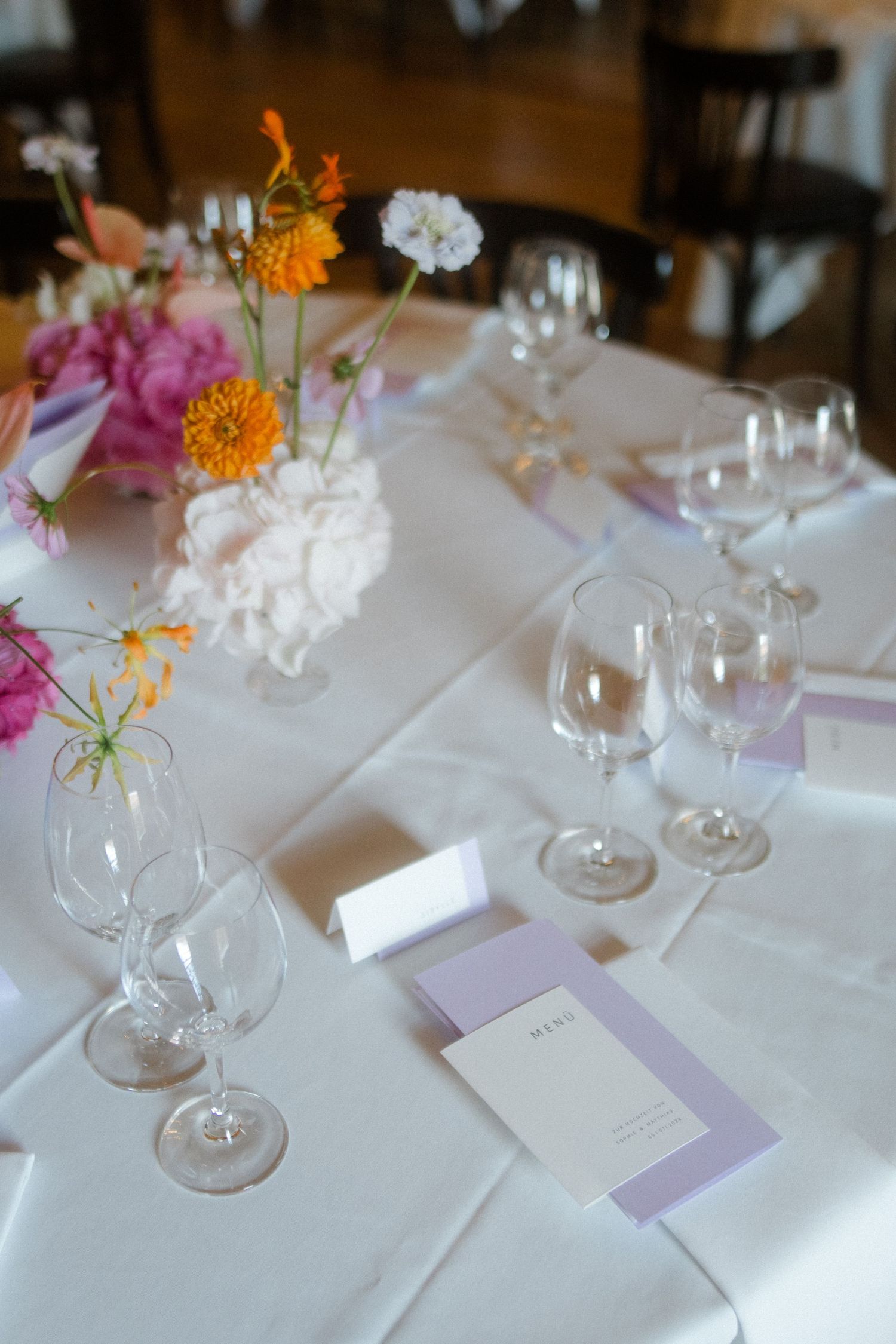 Elegant table setting with pink and orange flowers, wine glasses, and purple menu cards on white tablecloth.
