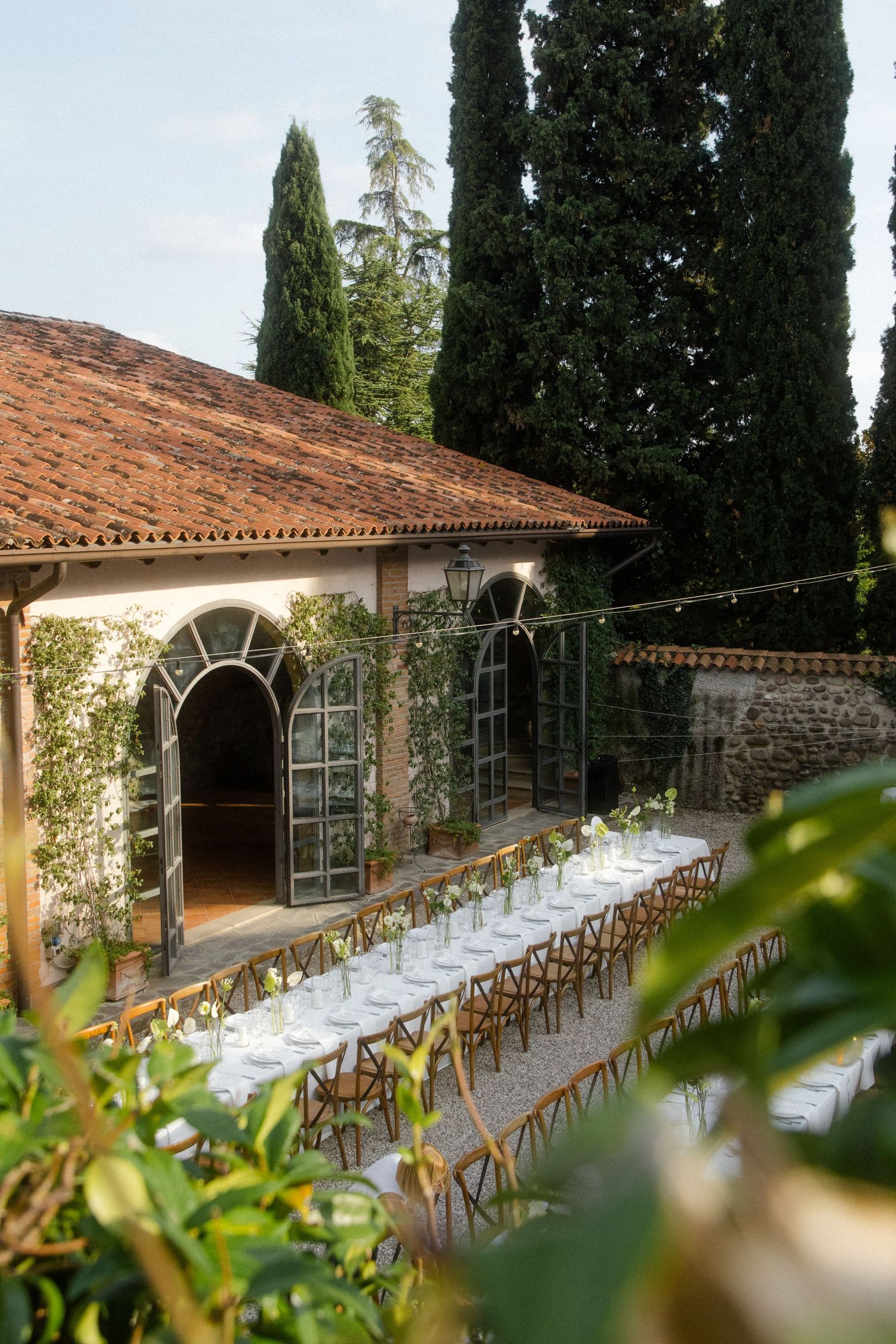 Long dining tables set up under rustic stone archway with cypress trees in background.
