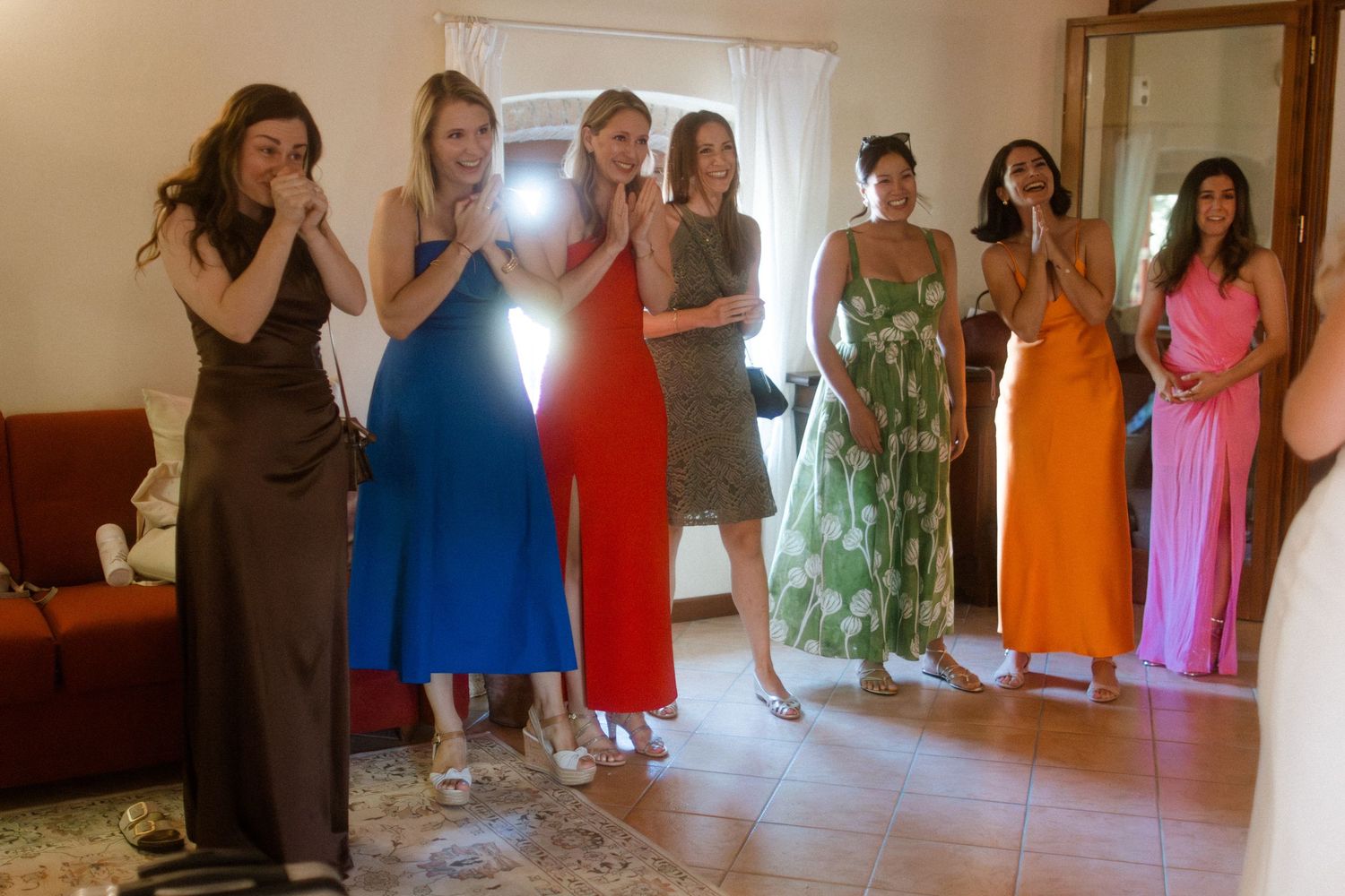 Wedding party members in vibrant dresses stand together in a sunlit room.