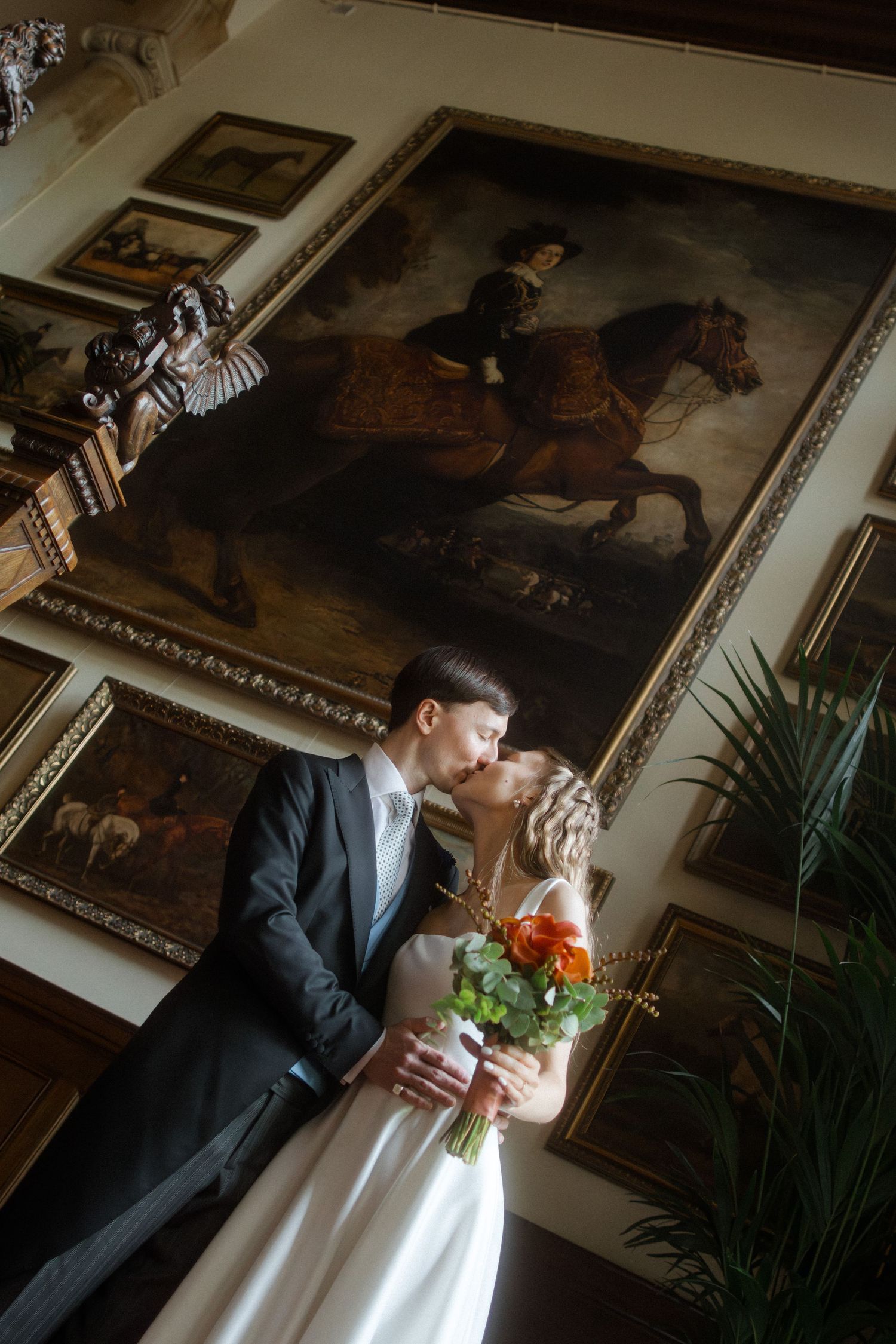 Wedding couple poses in elegant room with classical paintings and dark dramatic lighting.