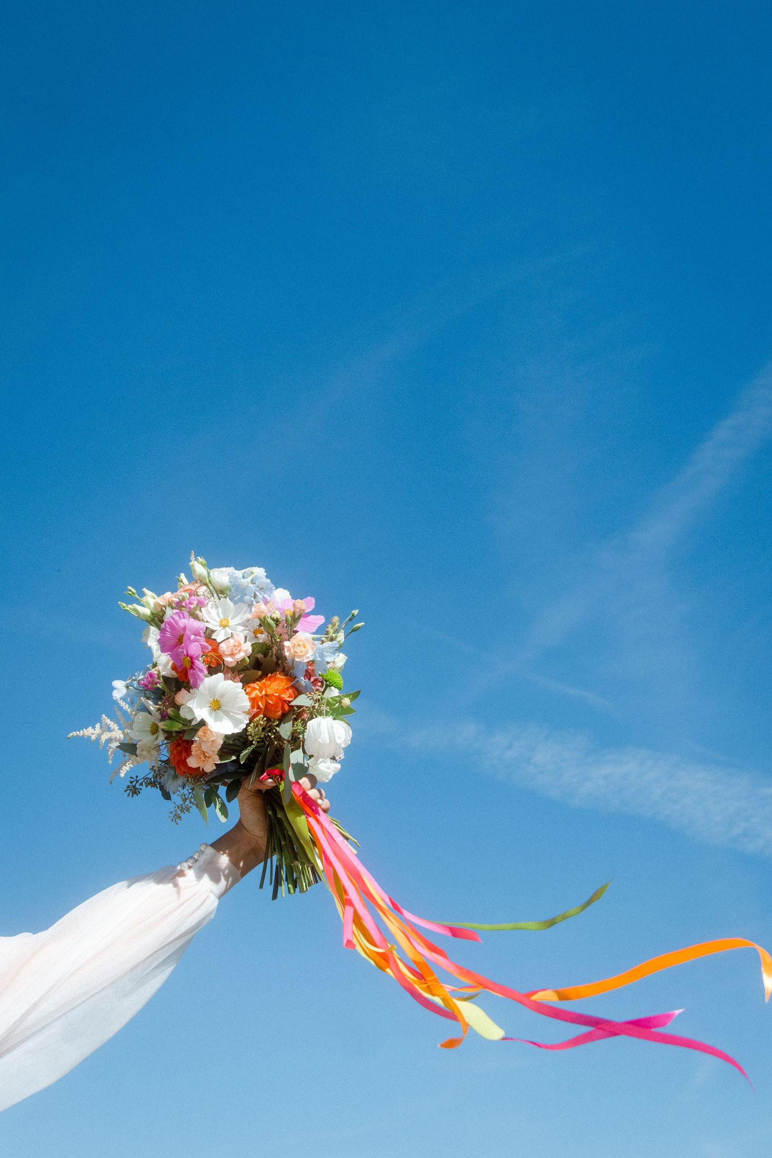 Colorful bridal bouquet with flowing ribbons against bright blue sky.