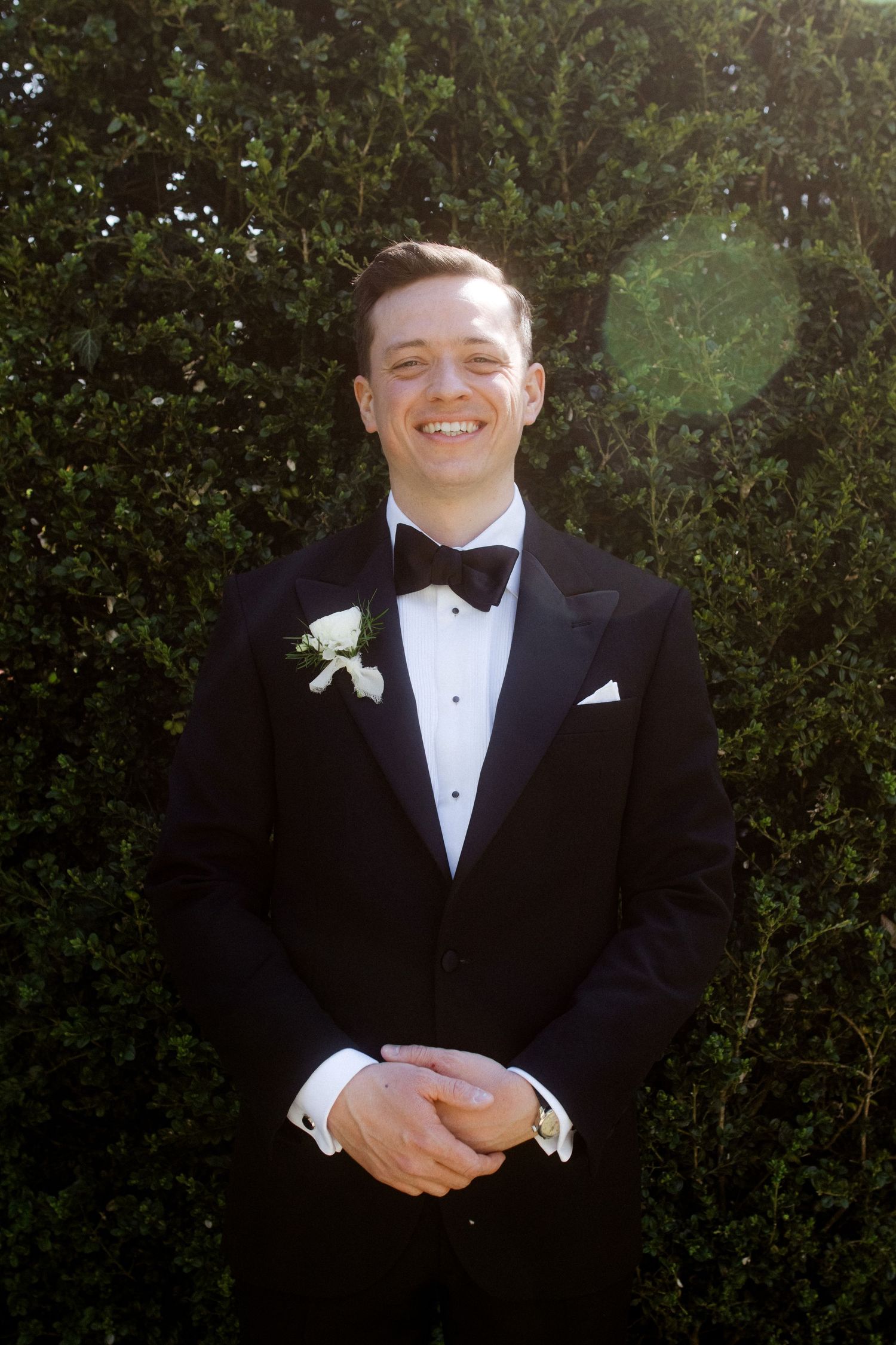 Smiling groom in black tuxedo with bow tie and white boutonniere standing before green hedges.
