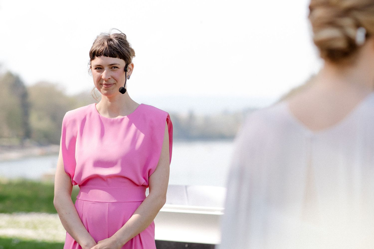 Woman in pink dress smiling at bride during outdoor wedding ceremony near a lake.
