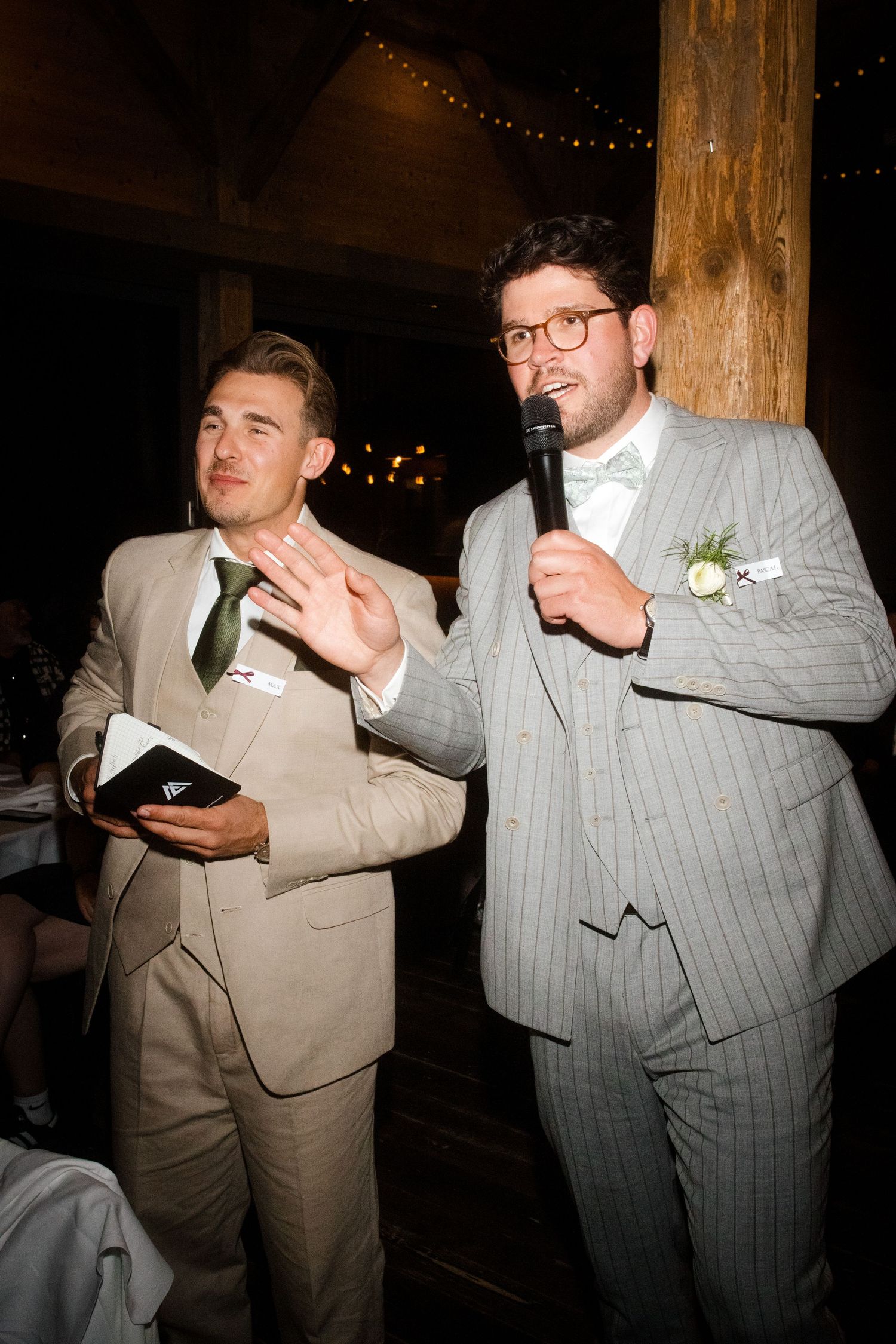 Two men in suits give a wedding speech, one holding a microphone and the other gesturing, in a rustic barn venue.