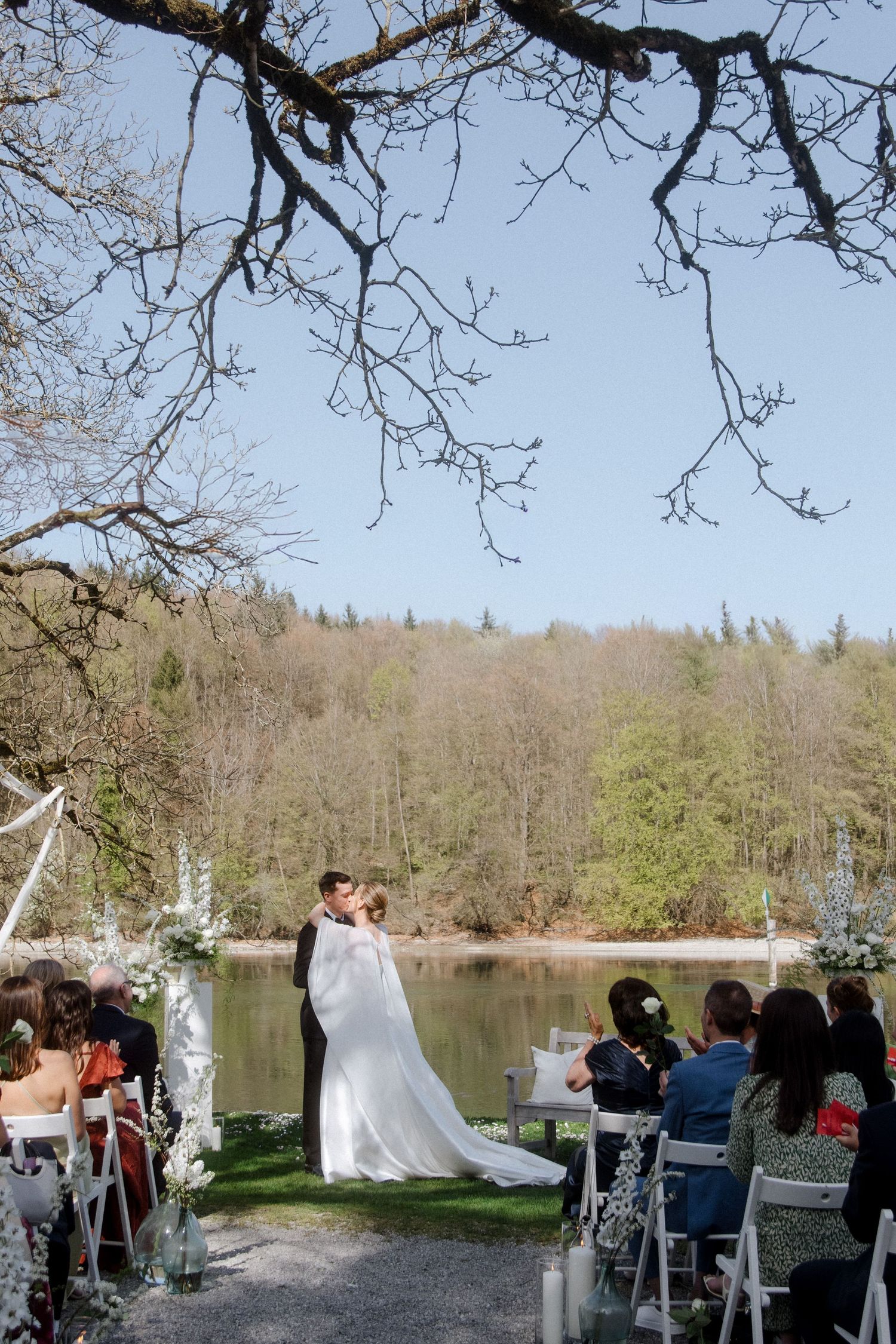 Bride and groom share first kiss at outdoor lakeside wedding ceremony surrounded by lush spring trees and white floral arch.