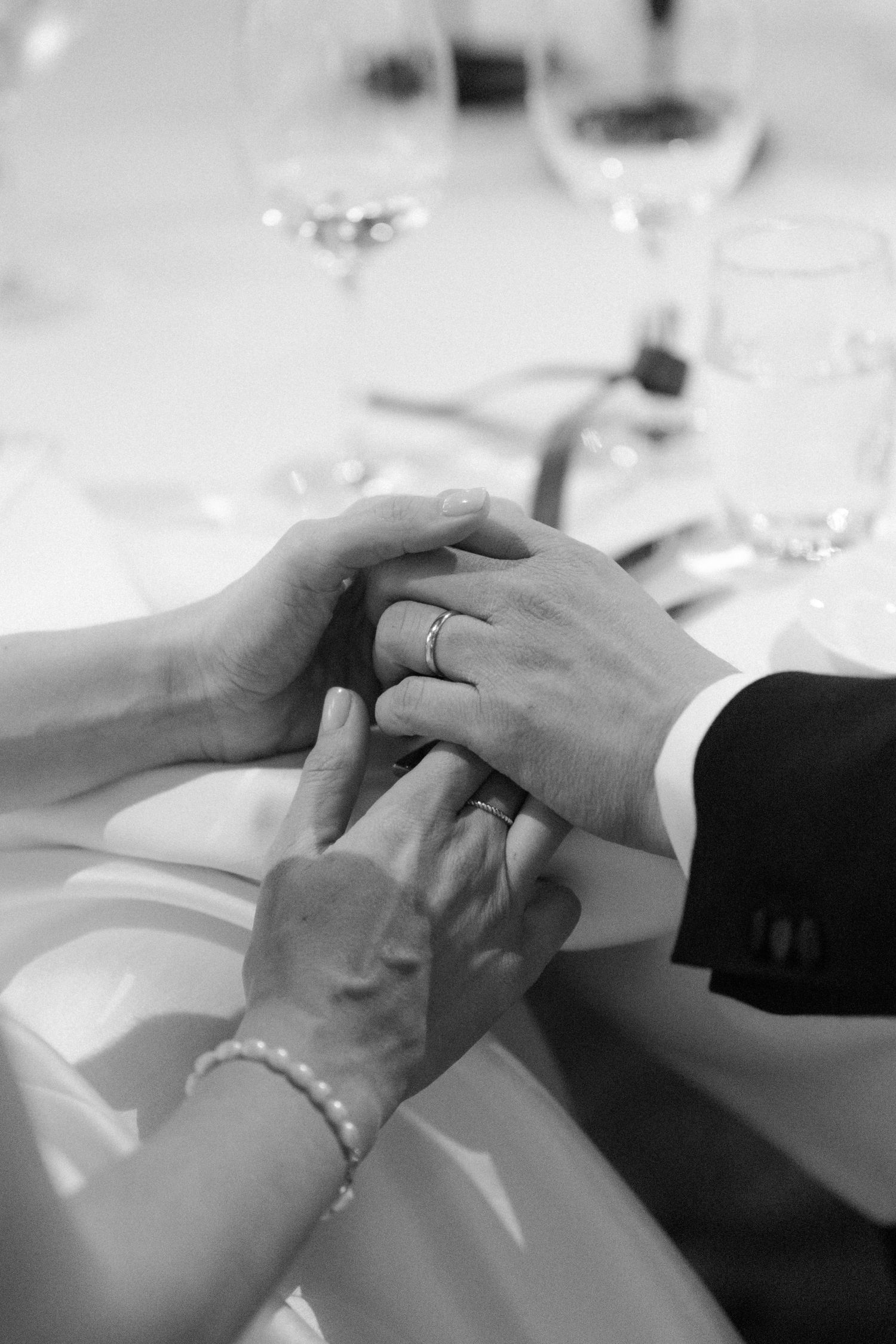 Black and white photo of newlyweds holding hands with wedding rings at reception table with wine glasses.