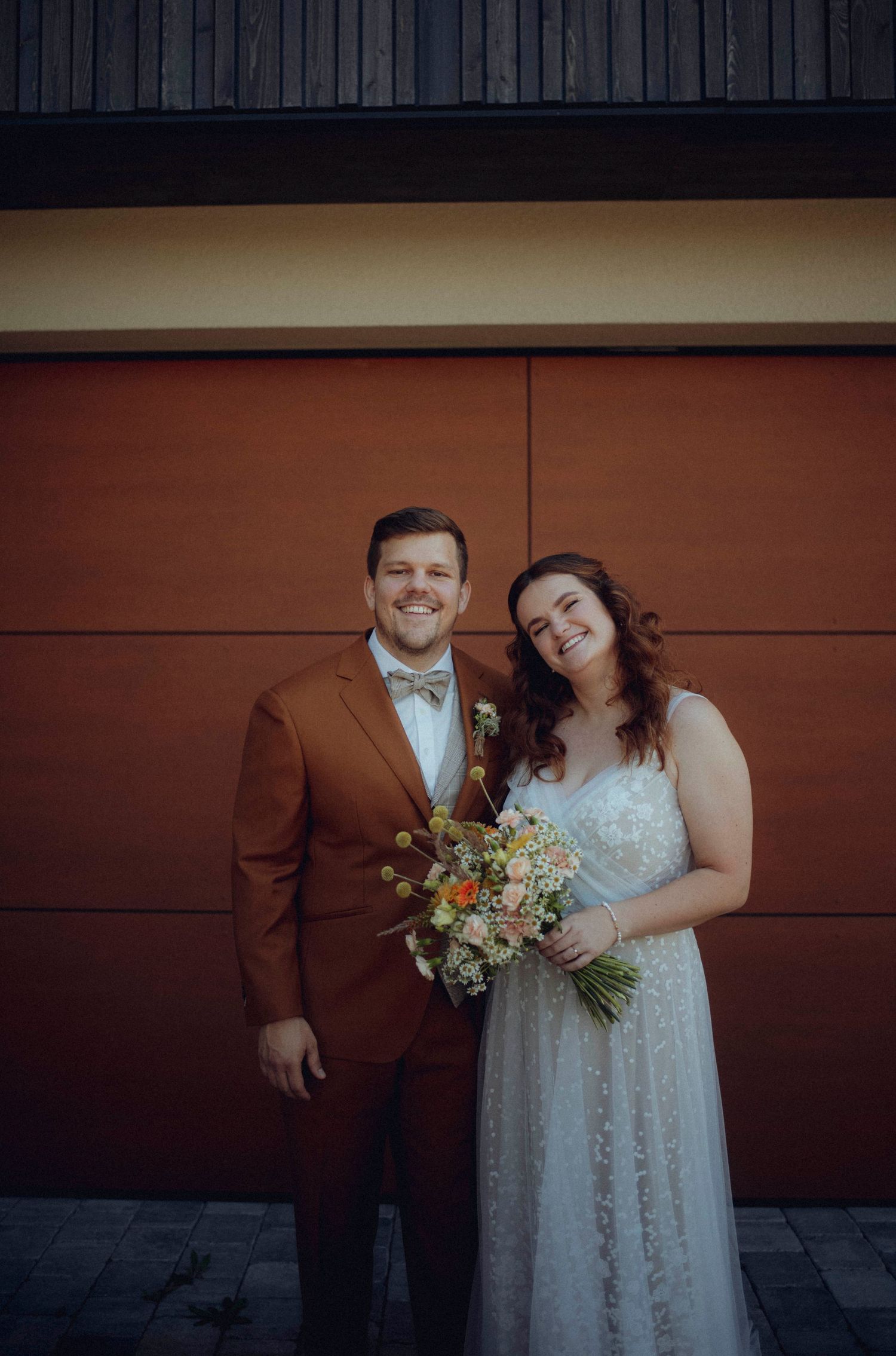 Wedding couple poses against brown wall in formal attire with autumn bouquet.